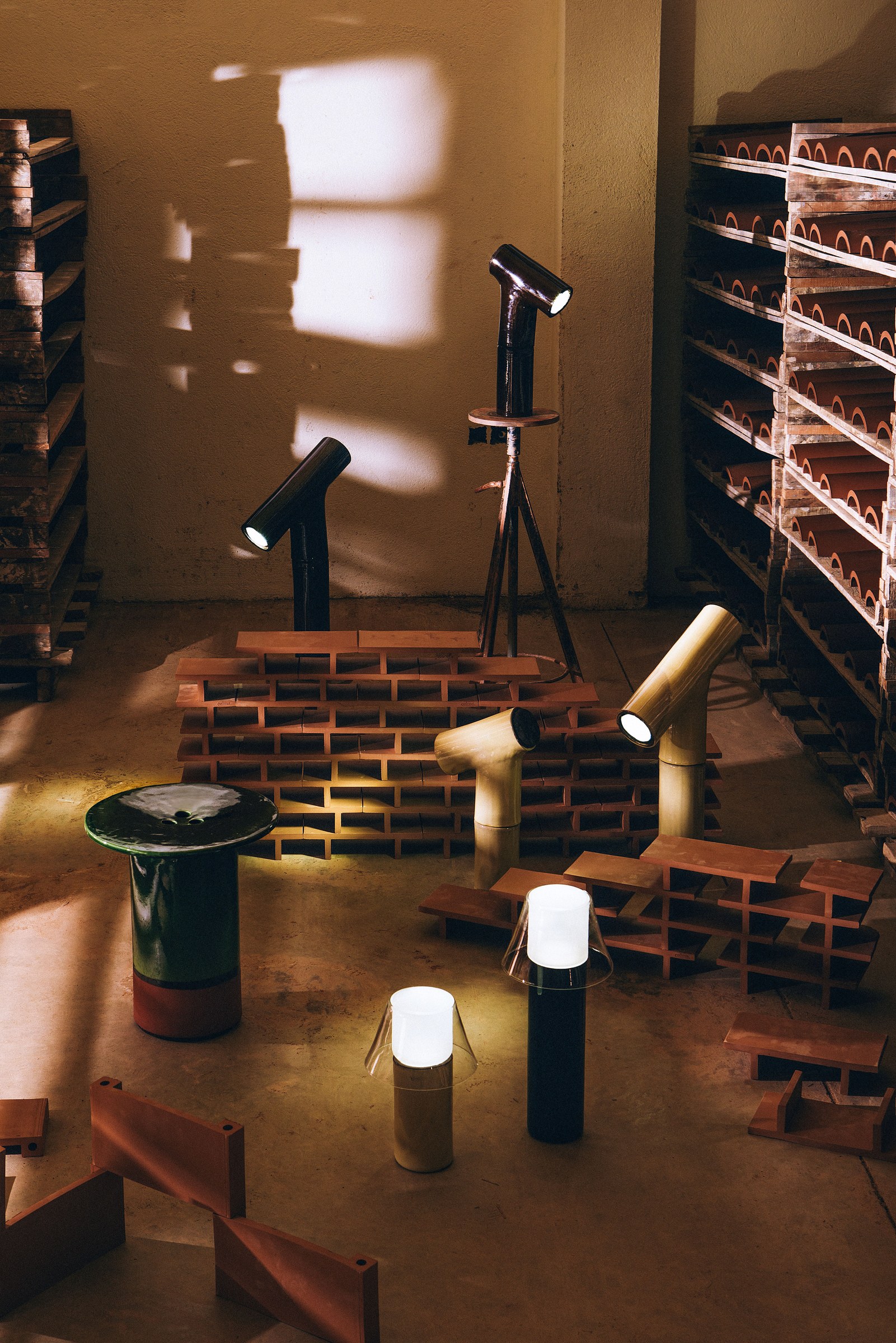 A green-glazed stool and several glazed ceramic lamps standing on the floor of a terracotta works, among stacked roof tiles and drying racks