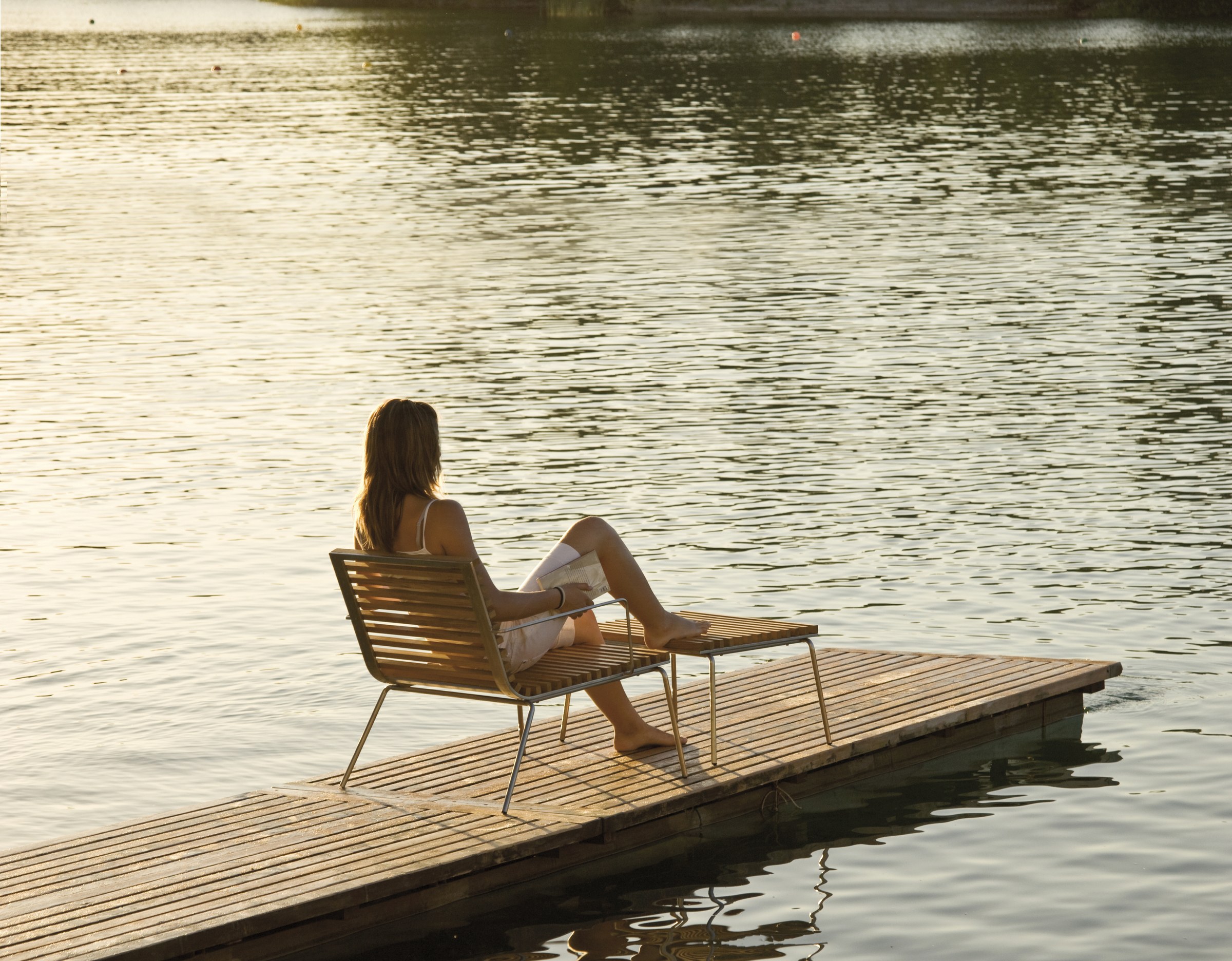 A woman sits side-on in a slatted Trama lounge chair with her feet up on its ottoman, reading, at the end of a timber jetty over a still lake at golden hour, a wooded far shore beyond.