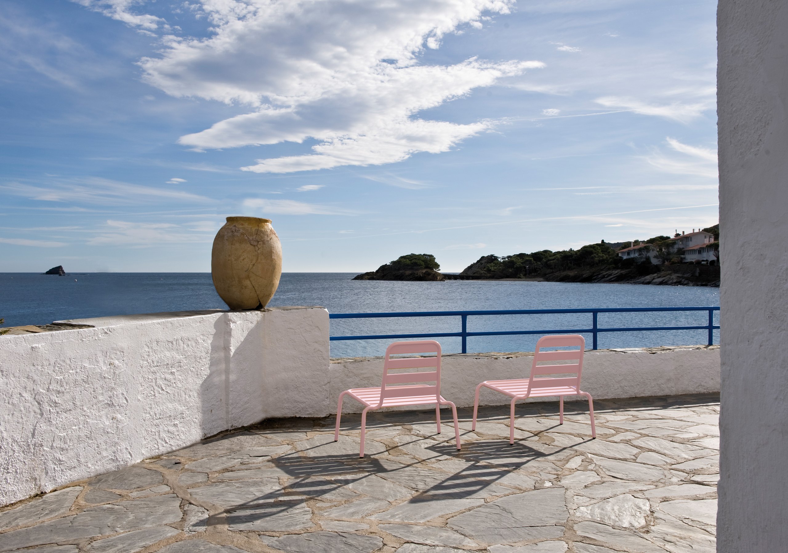 Two pale-pink lounge chairs seen from behind on a whitewashed stone terrace, a terracotta urn on the parapet beside them, looking over a blue rail to a Mediterranean cove with a pine headland and white villas