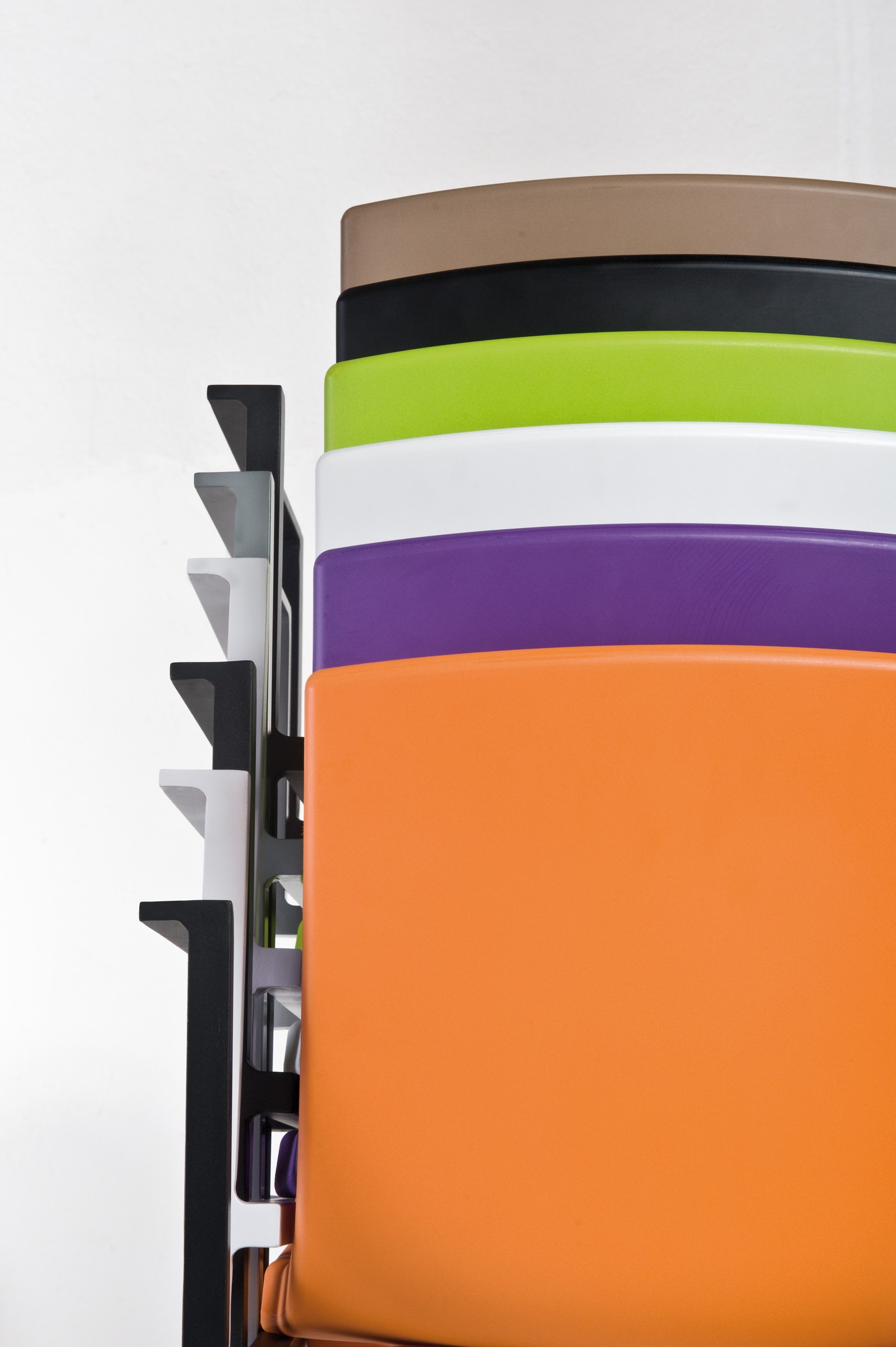 A stack of moulded chair seats seen from behind — orange, violet, white, lime, black and taupe — nested on their frames against a white wall