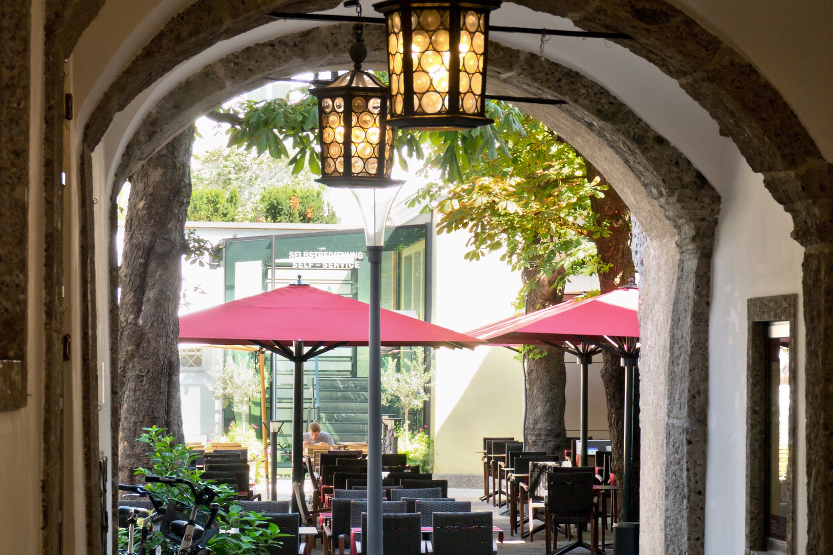 Two Big Ben umbrellas with red square canopies on black frames, seen through a stone archway hung with lanterns.