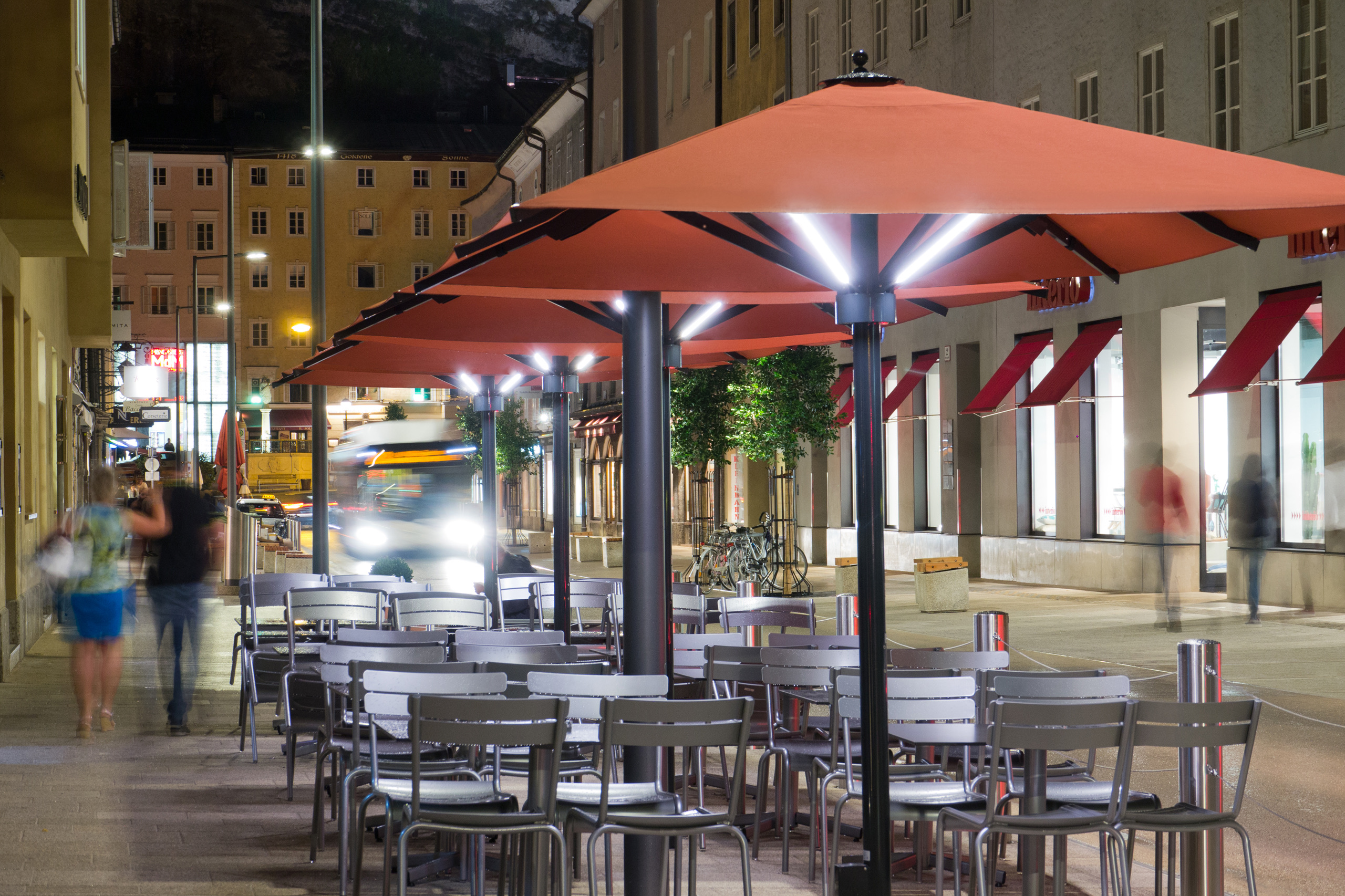 Caravita Big Ben umbrellas at night: square orange canopies lit along their struts above a row of café tables.