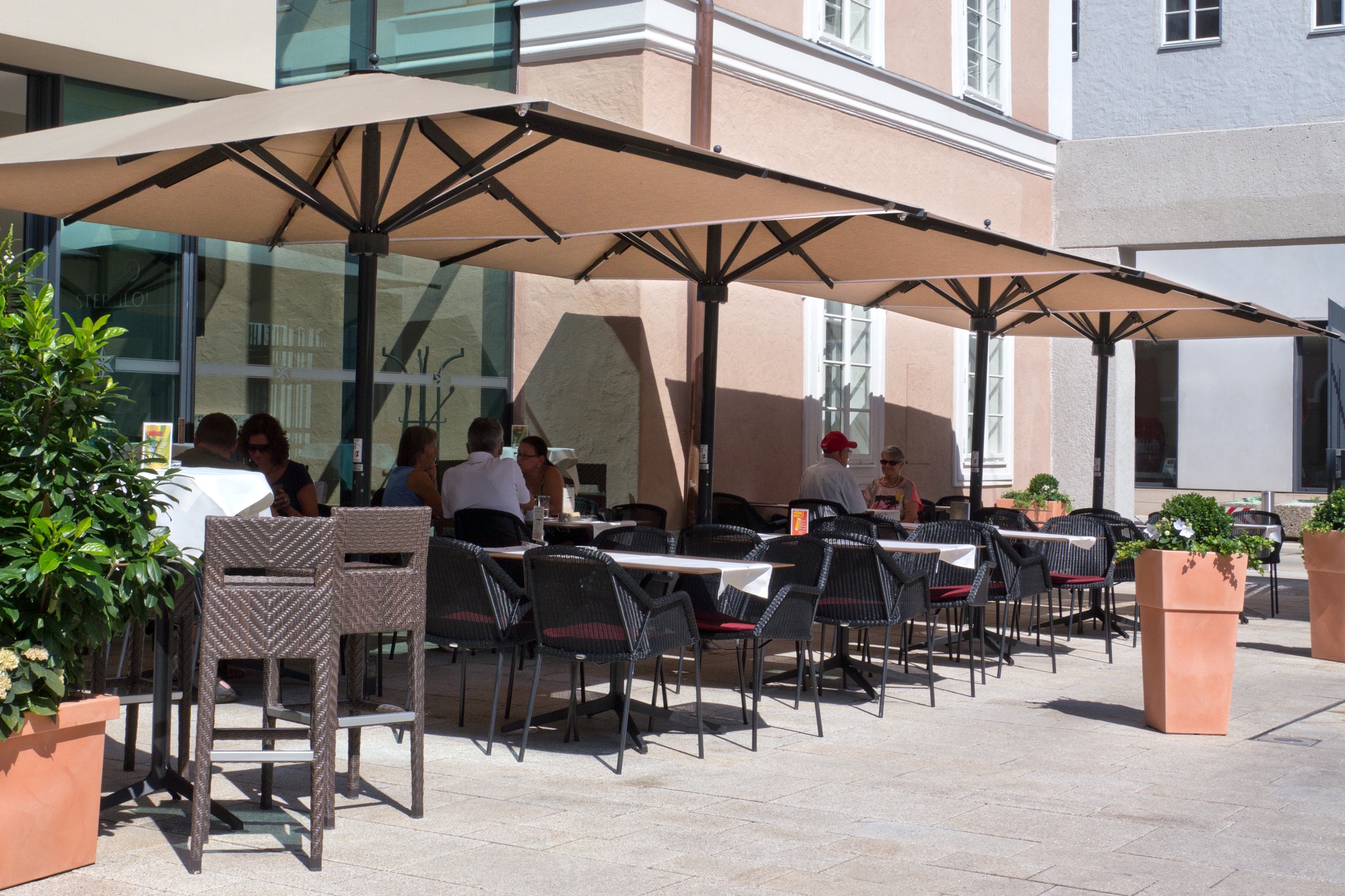 Big Ben umbrellas with square natural-coloured canopies on brown frames over a row of café tables in a paved square.