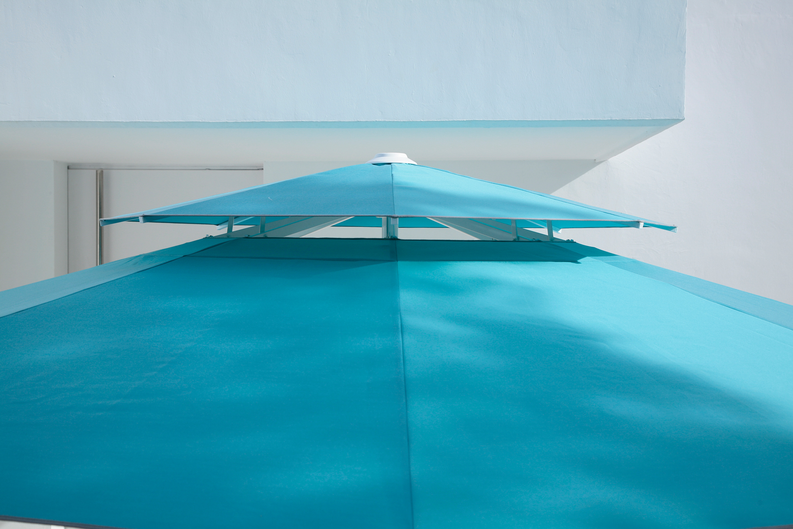 An aqua Caravita canopy with its raised wind roof, seen from below against a white wall.