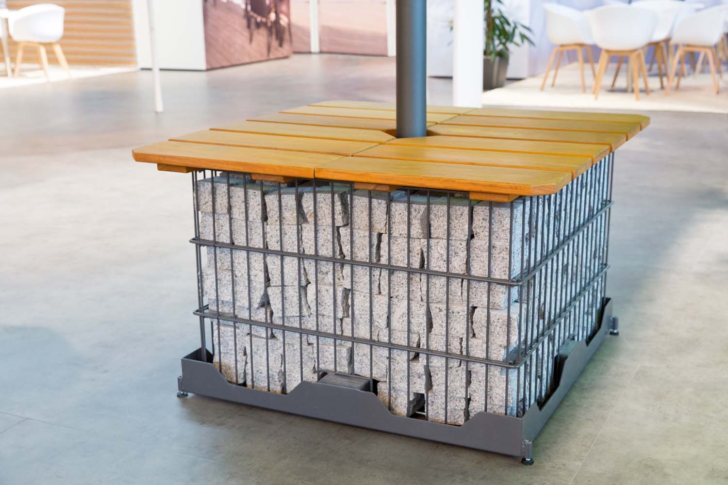 A steel gabion base filled with stone under a light timber slatted top, an umbrella pole rising through its centre.