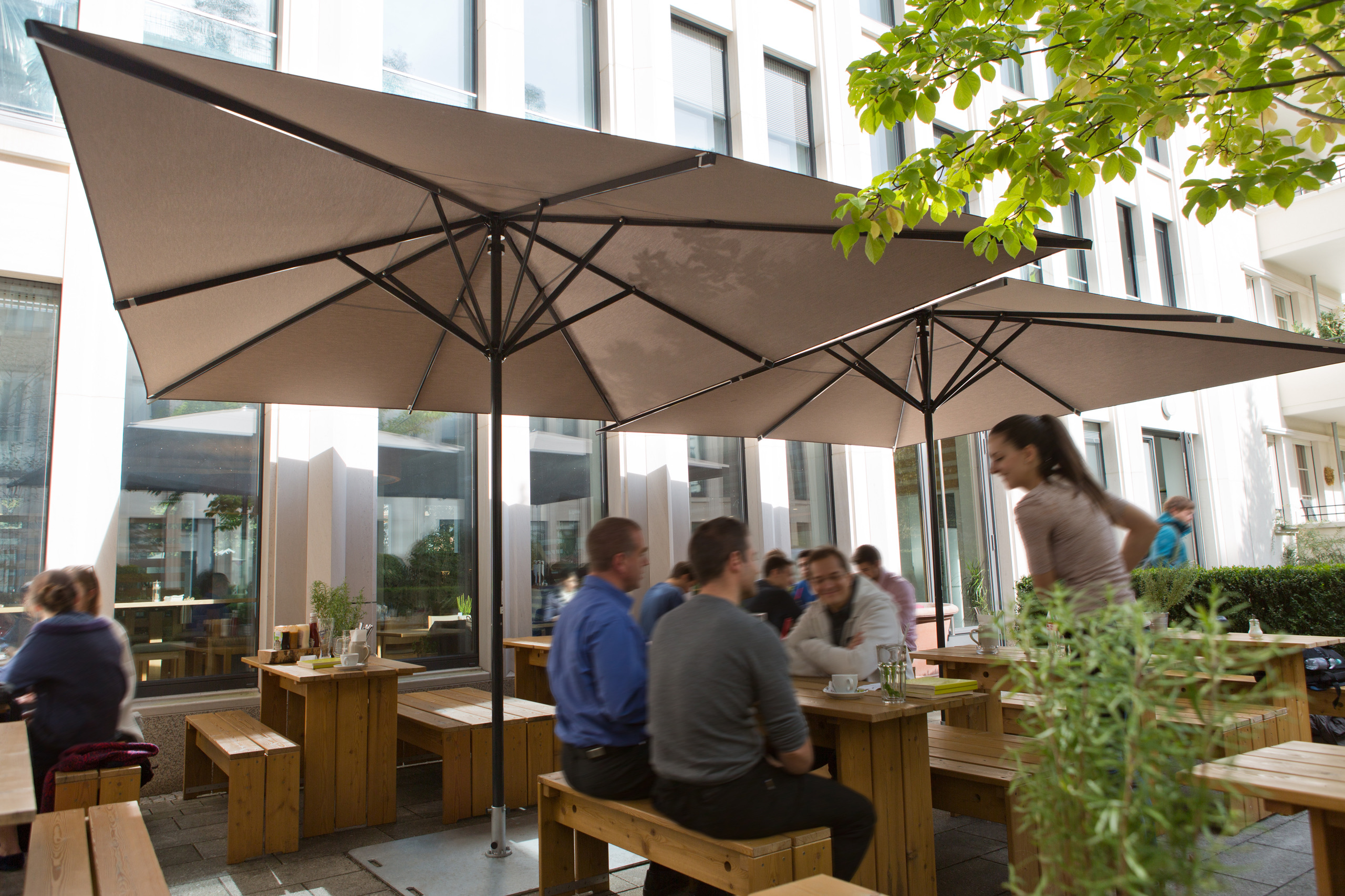 Samara umbrellas with square cigar-coloured canopies on black frames over a busy outdoor lunch.