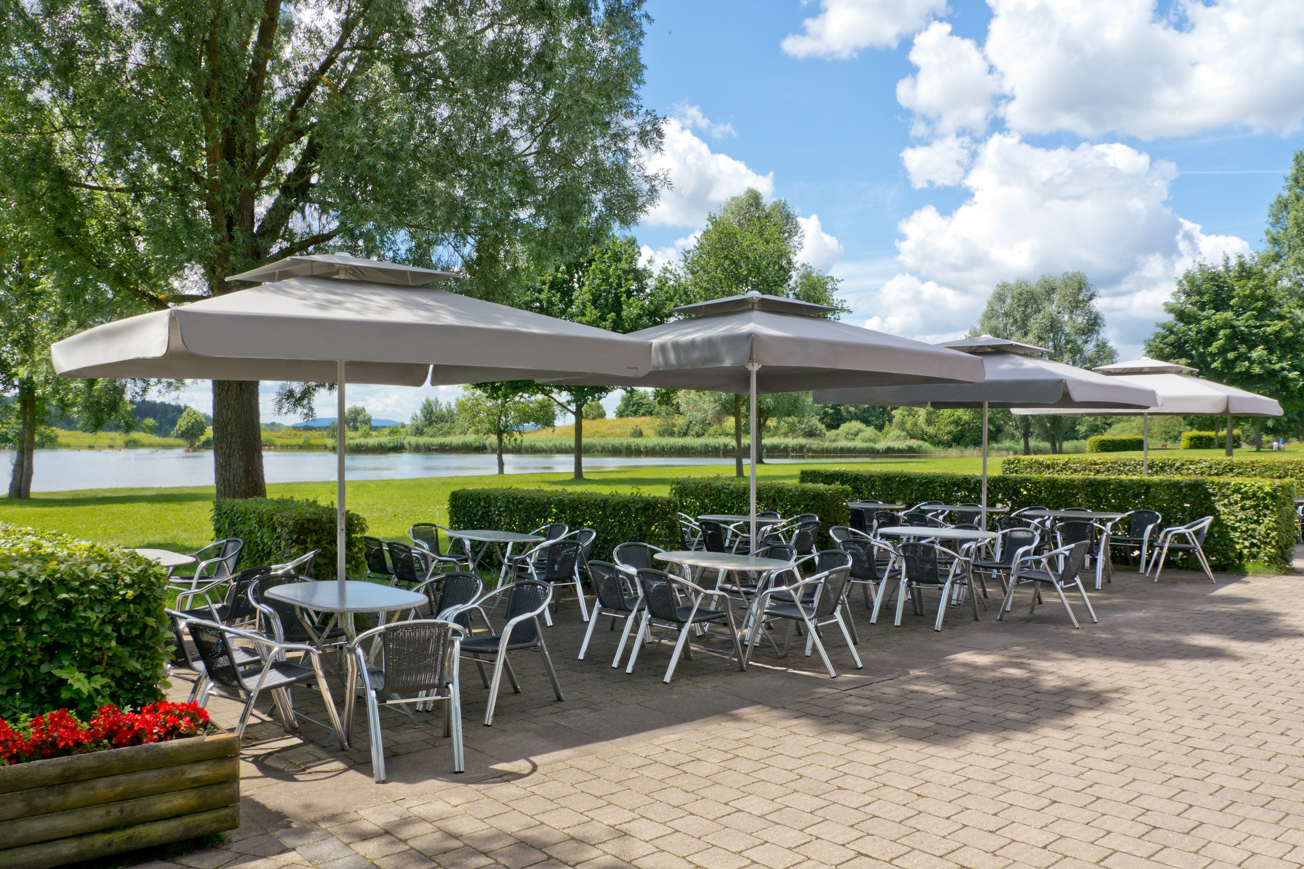 A row of Samara umbrellas with grey canopies, valances and wind roofs over restaurant tables beside a lake.