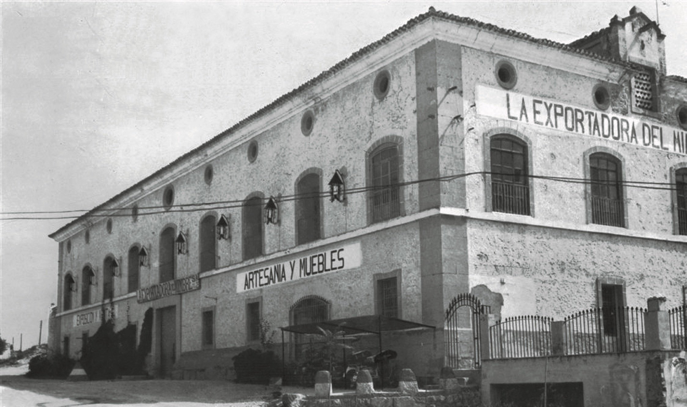 A weathered stone warehouse on a hillside corner, its facade lettered 'La Exportadora del Mimbre — Artesanía y Muebles', arched windows along the upper storey and a wrought-iron gate below.