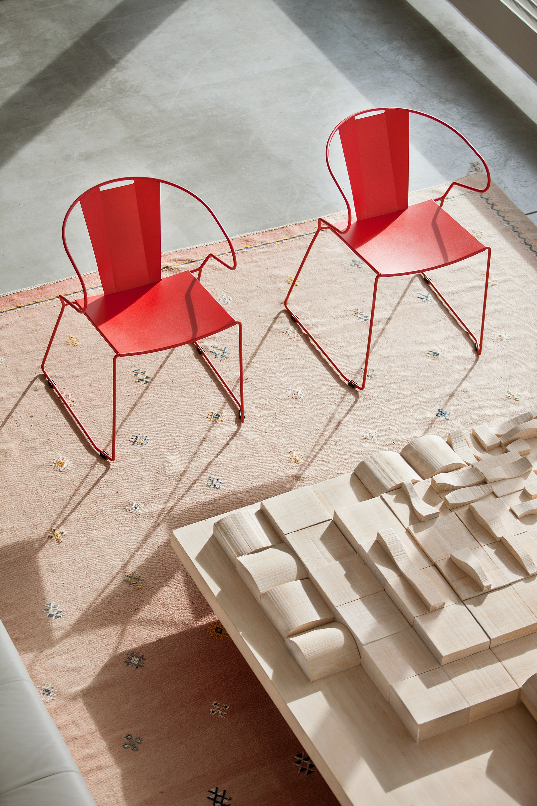 Two red steel chairs on a pale rug, casting long shadows.