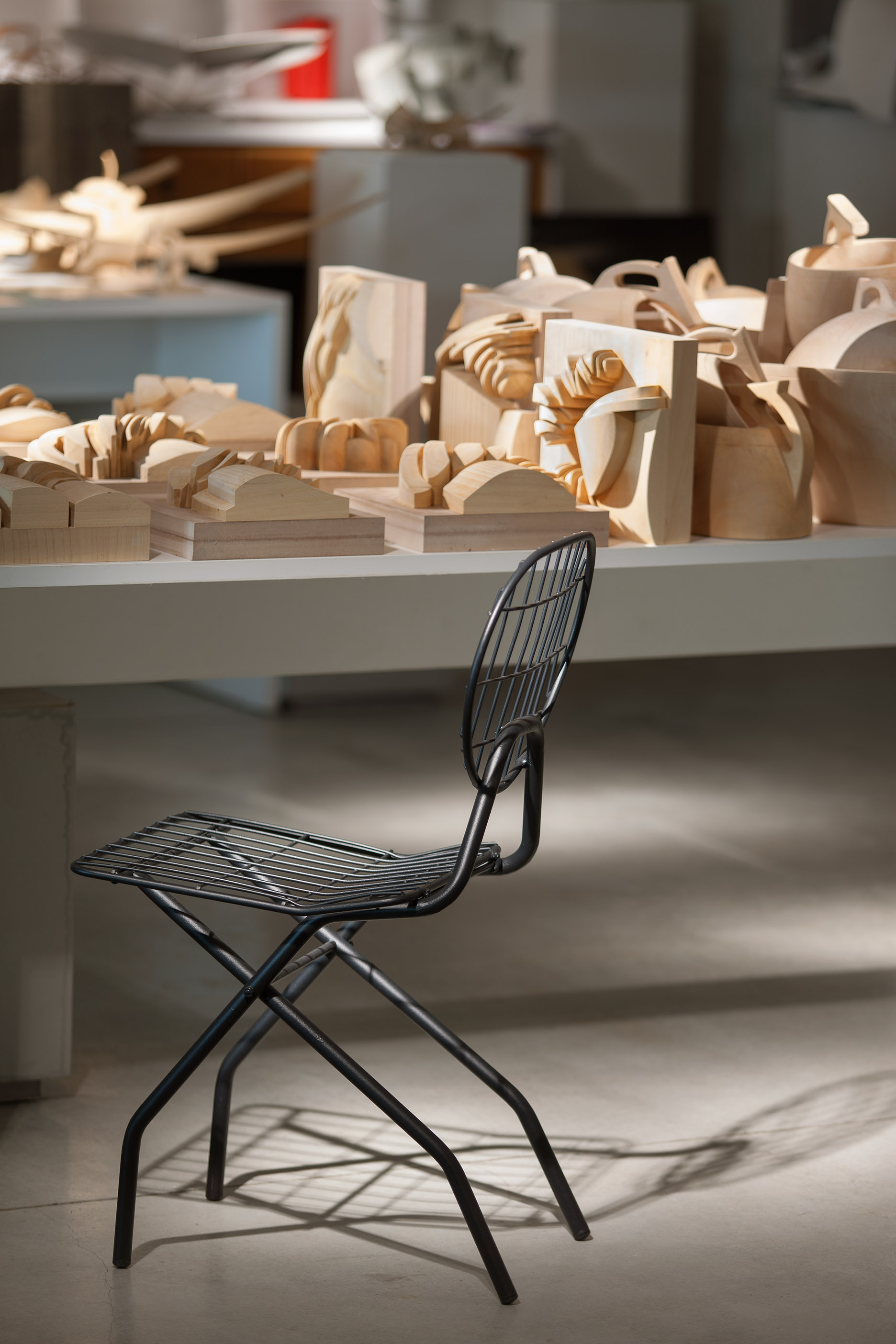 A black wire folding chair standing in front of a workbench stacked with raw timber study blocks