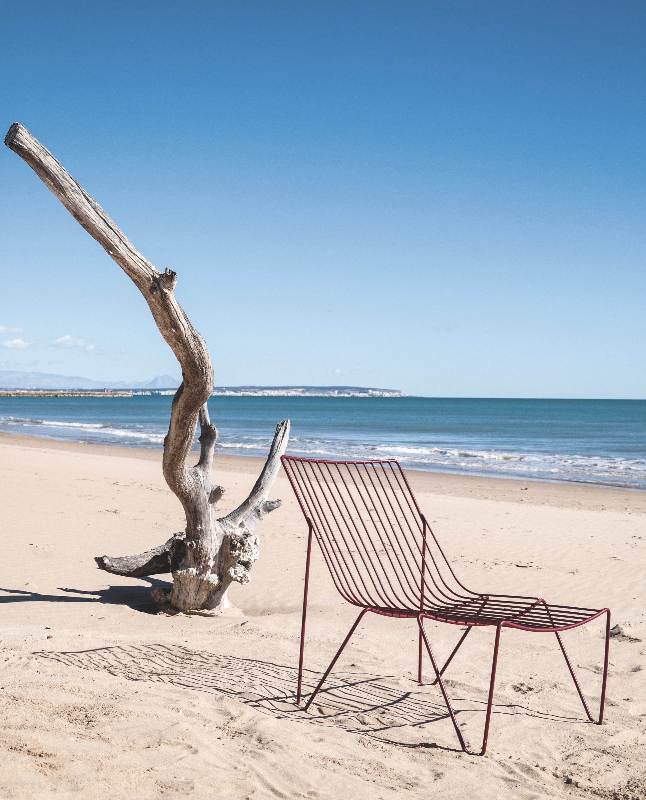 A burgundy galvanized-wire lounge chair on open white sand beside a bleached driftwood spar, the sea behind.