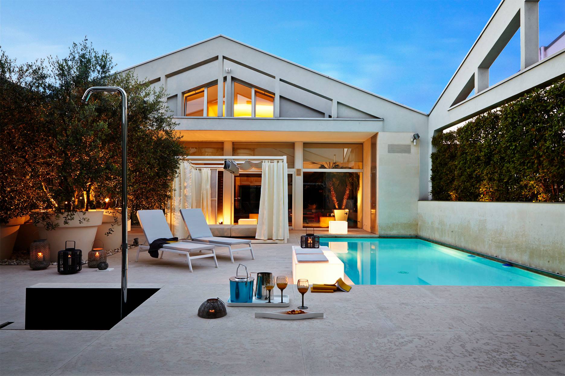 A white-cushioned Victor sunlounge on an illuminated terrace at dusk, warm light spilling from a timber-and-glass pavilion behind, lanterns and drinks laid out beside the pool.