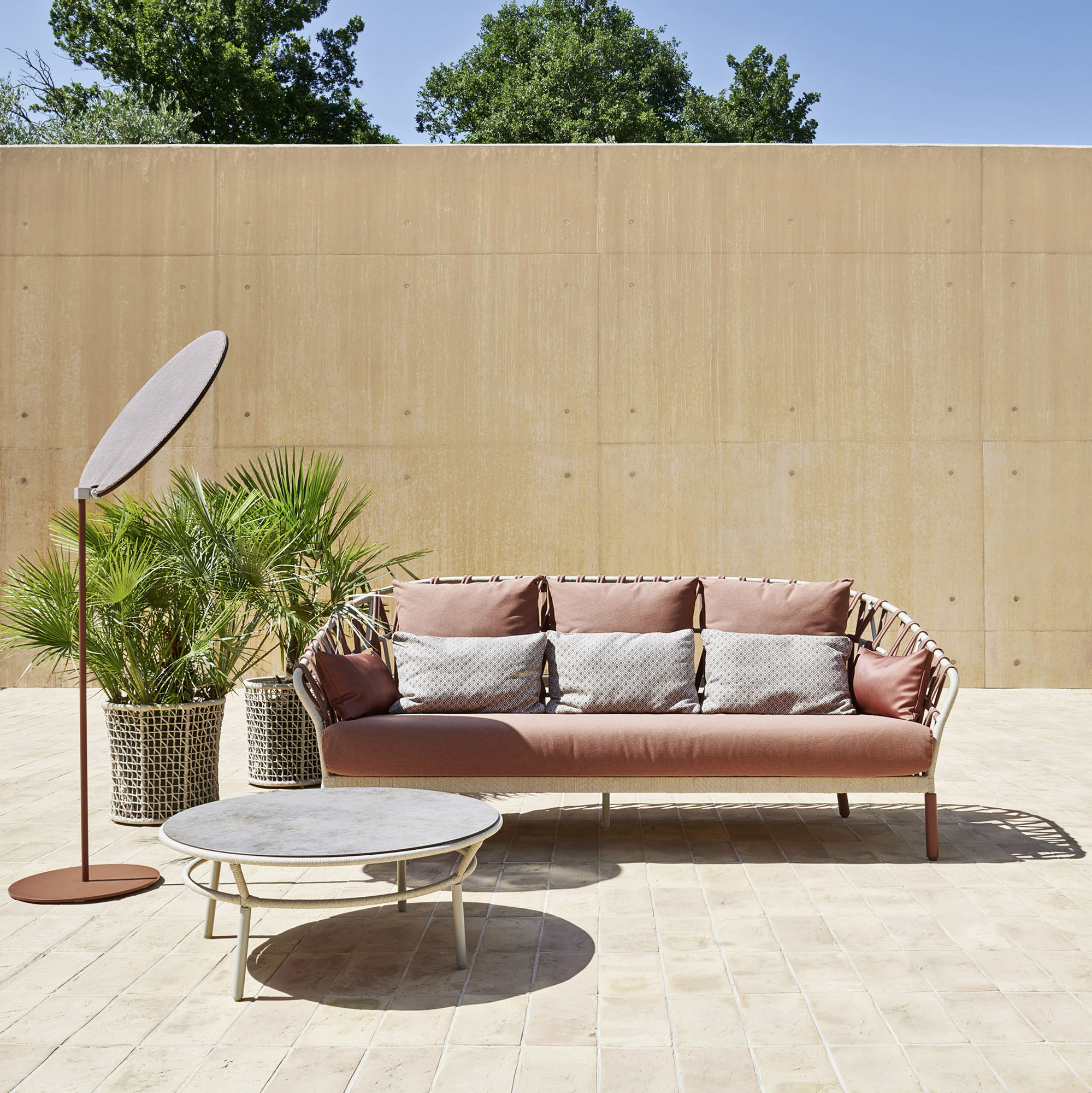 A woven sofa in terracotta tones with a tilting shade, on a paved terrace.