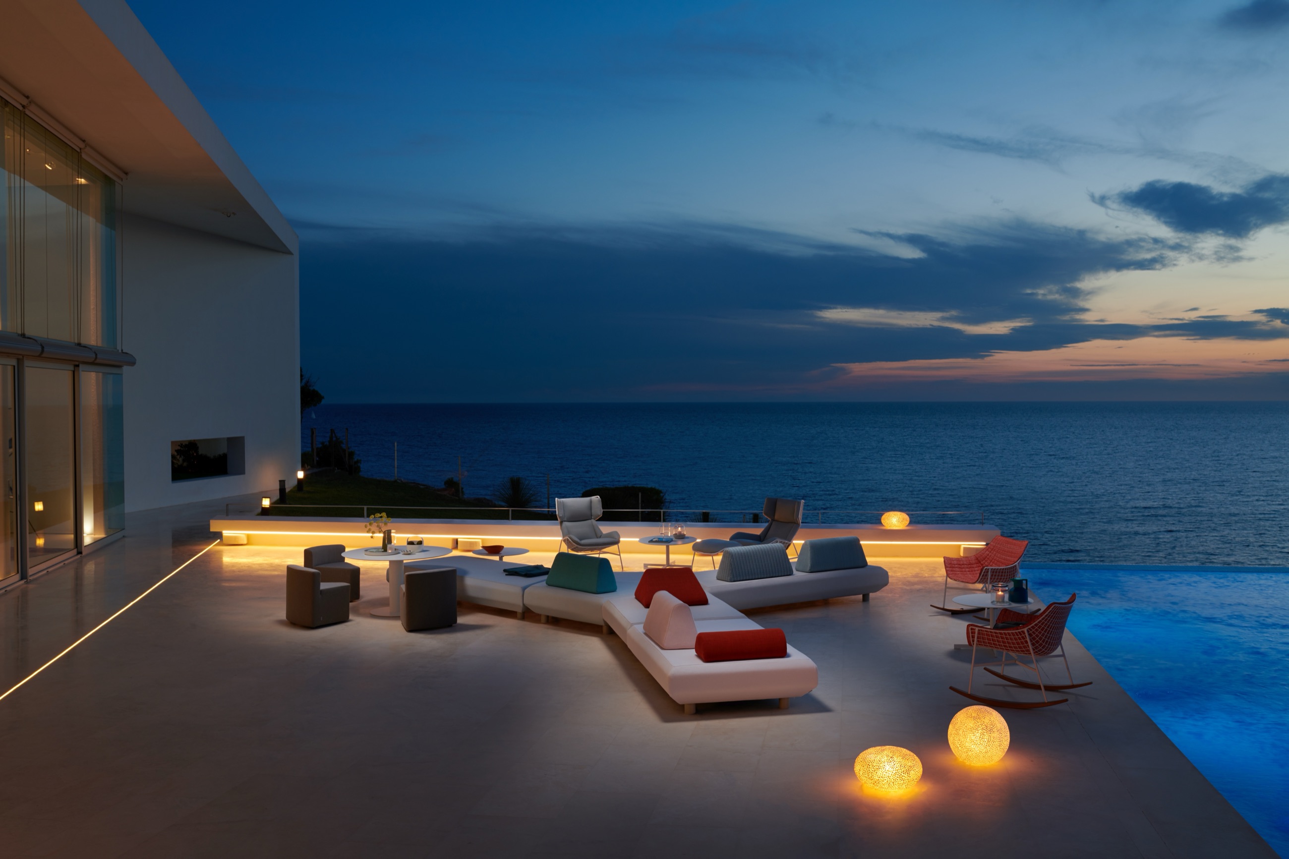 A terrace at blue hour above the sea: low modular seating and sunloungers on pale stone, a lit infinity pool to one side.