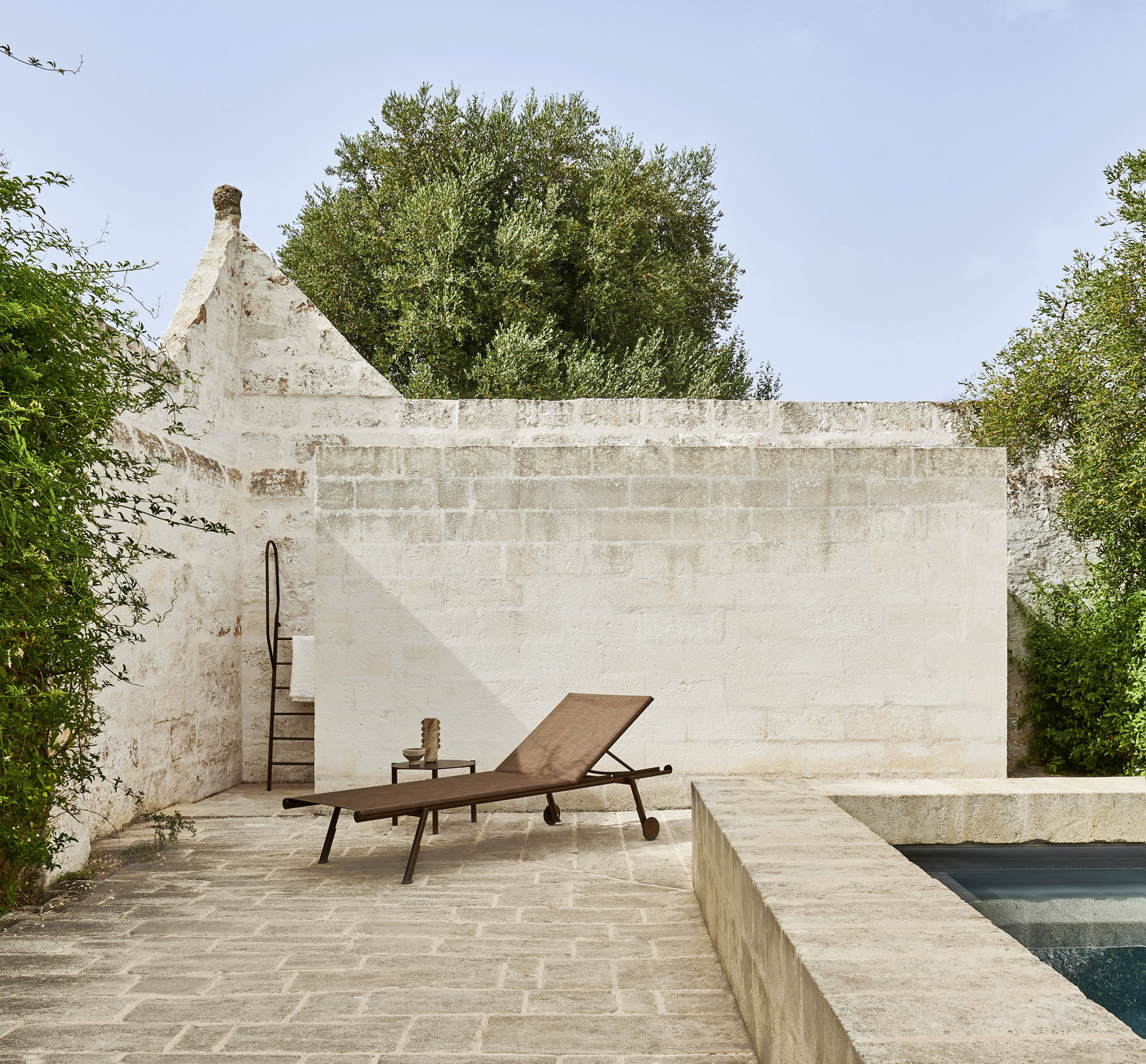 A bronze-framed Bahia sling sunlounge and round side table on a sunlit limestone courtyard, a conical stone roofline and olive canopy above, a plunge pool at the corner.