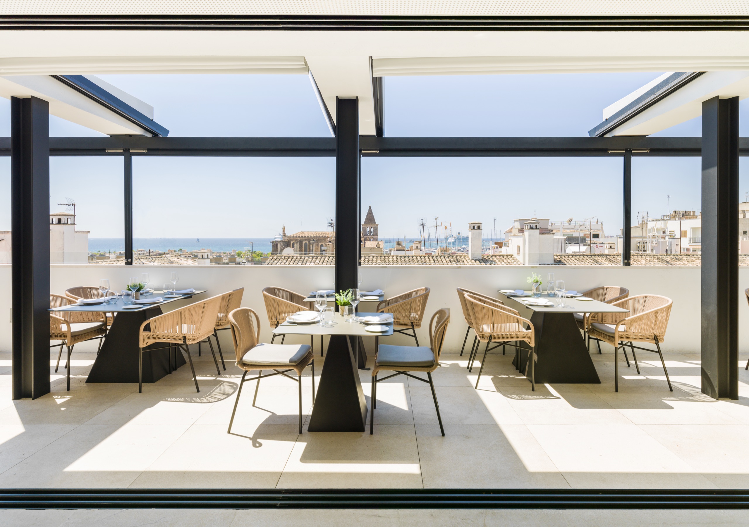 A rooftop restaurant terrace under a retractable pergola, laid with dark pyramid-base dining tables and woven dining chairs, overlooking a coastal town's terracotta rooftops, a bell tower and the sea beyond