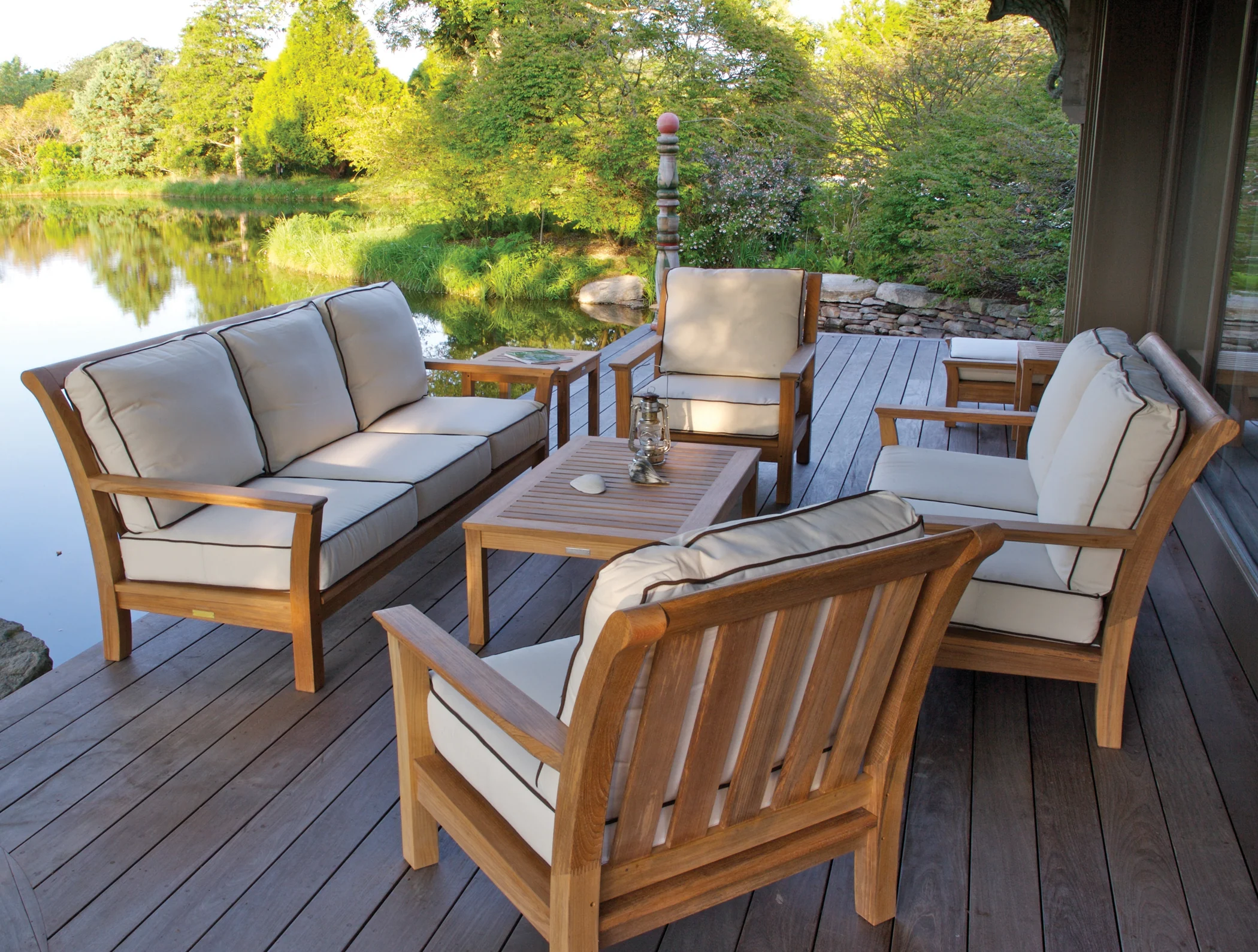 A teak deep-seating group with cream cushions on a weathered deck at the edge of a still pond
