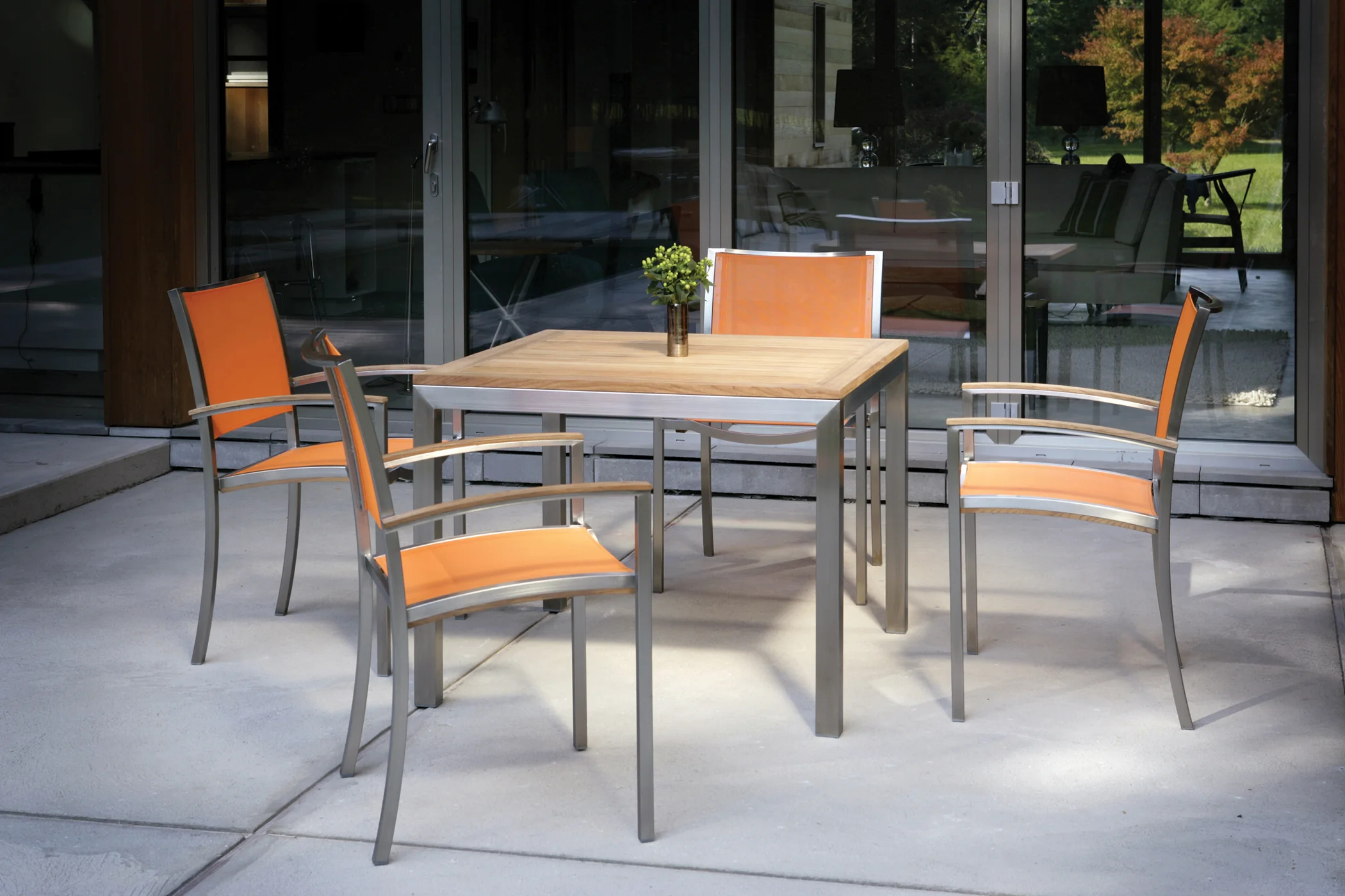Four stainless and teak dining armchairs in orange sling around a square teak table on a terrace outside a glazed house