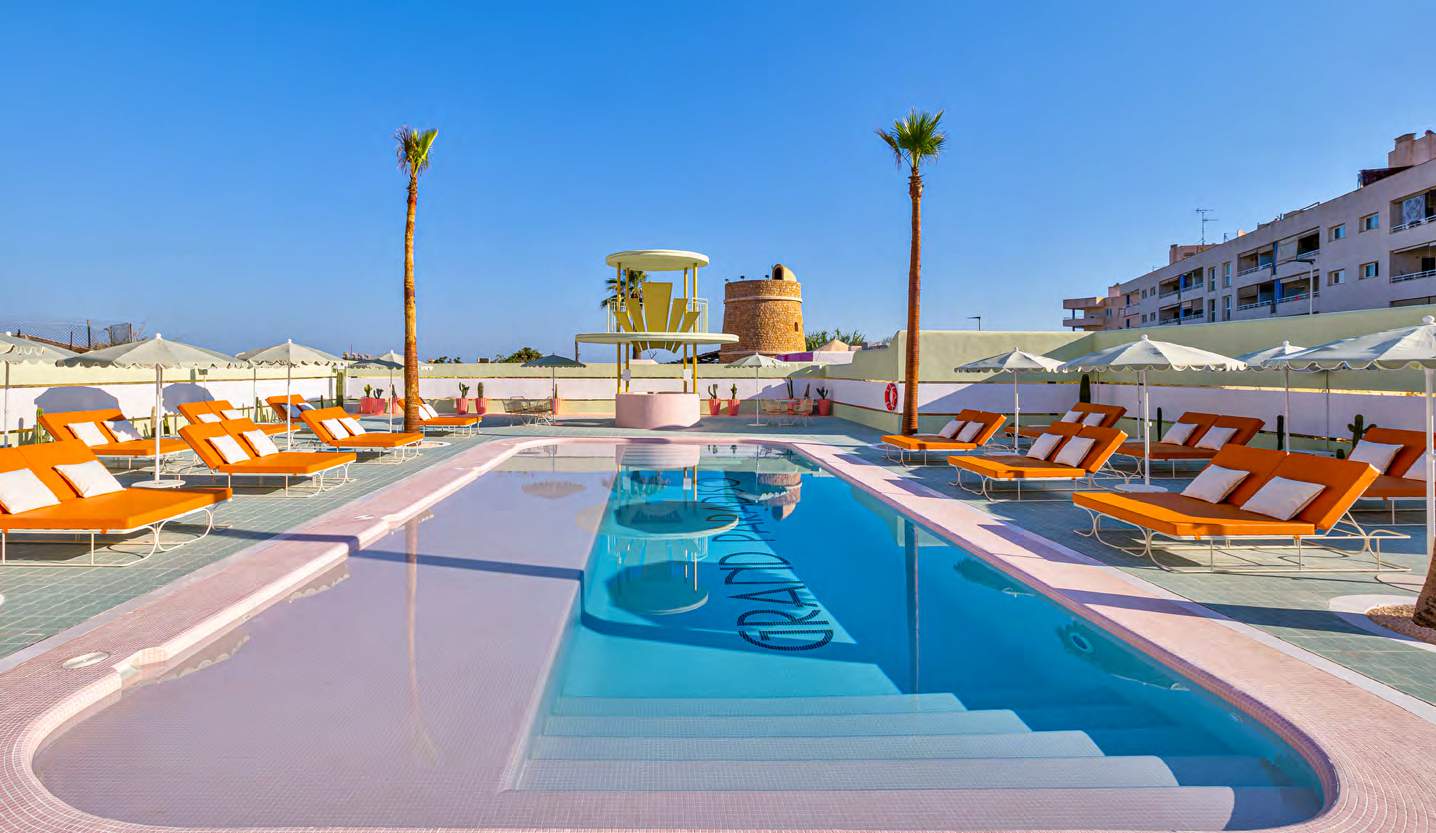 The pool at Grand Paradiso Ibiza in full sun, GRAND PARADISO lettered across the pool floor, rows of burnt-orange sunloungers under scalloped cream parasols on both terraces, a lemon-yellow bar pavilion and a stone watchtower behind.
