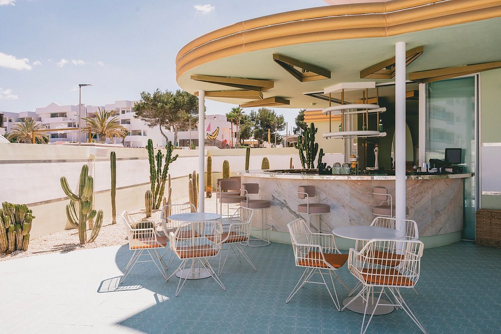 The bar pavilion seen across its terrace: a pale marble counter under a ribbed lemon-yellow canopy, white wire armchairs around white round tables on mint tiling, cacti along a low white wall and the town behind.