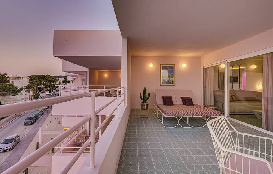 A guest room opening onto a white balcony above pines and the coast road, a daybed inside carrying pink-and-burgundy striped cushions and a white wire chair at the threshold.