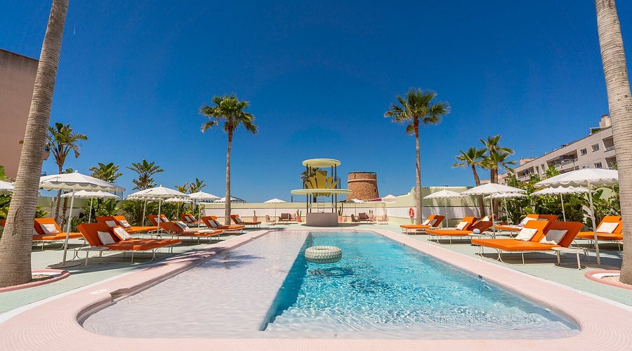 The same axis at midday under a deep blue sky, the stone watchtower and the yellow pavilion centred between palms, loungers and cream parasols down both sides of the water.