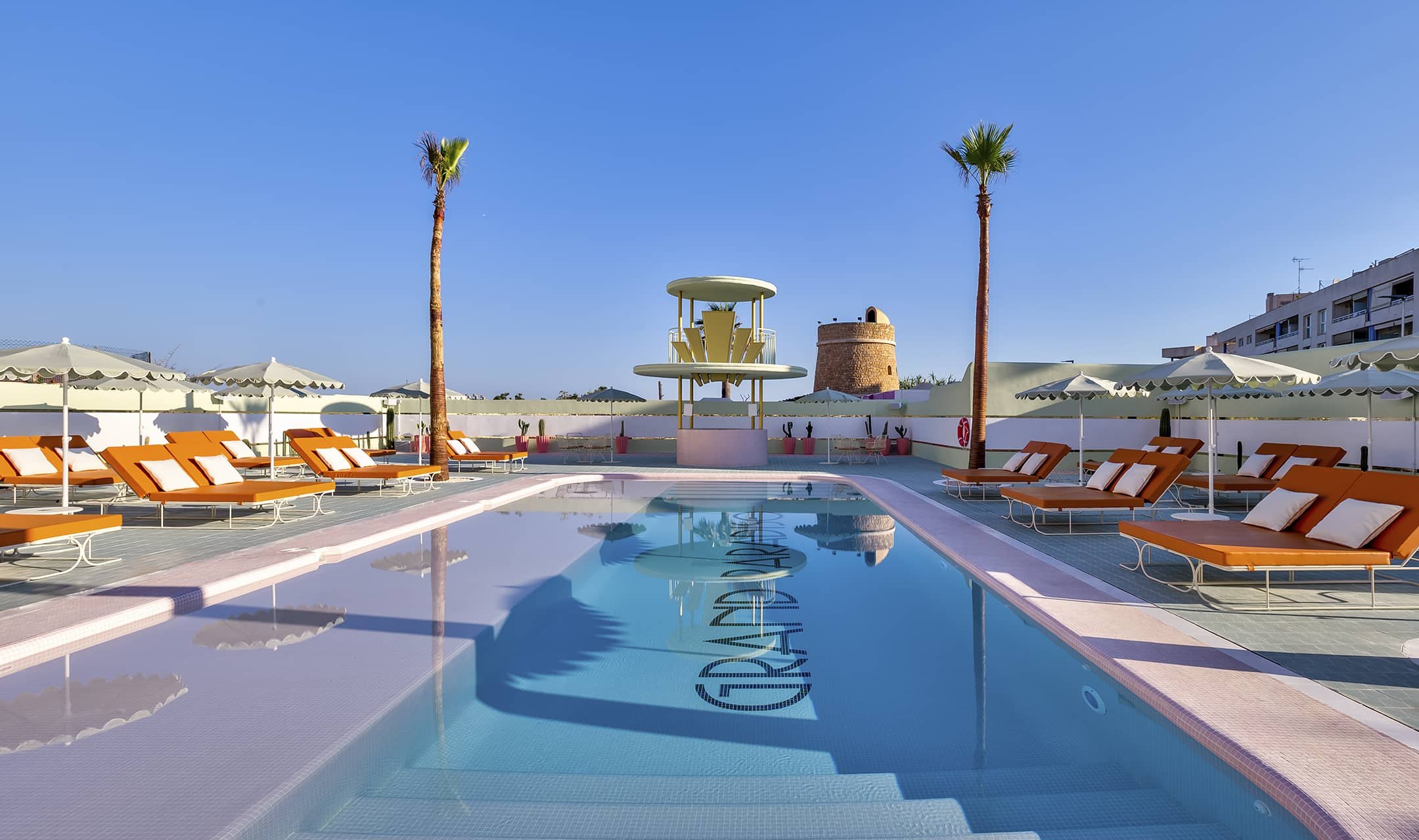 The pool on its long axis at golden hour, palms either side of the lemon-yellow bar pavilion, burnt-orange loungers under cream parasols down both edges and GRAND PARADISO lettered across the pool floor.