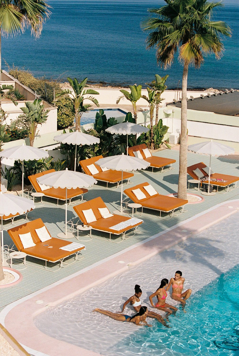 The poolside from above the terrace: burnt-orange loungers under white parasols set around a small pool, swimmers on the entry steps, palms and the sea beyond.
