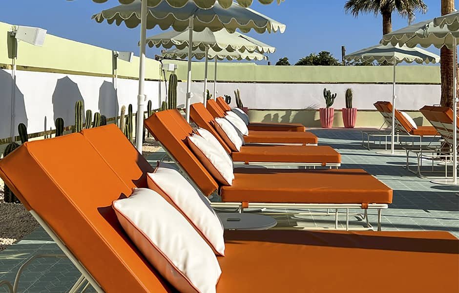 A close row of double sunloungers with white tubular frames, burnt-orange mattresses and white bolster cushions, on mint terrazzo under scalloped green-and-cream parasols, with cacti planted between them.