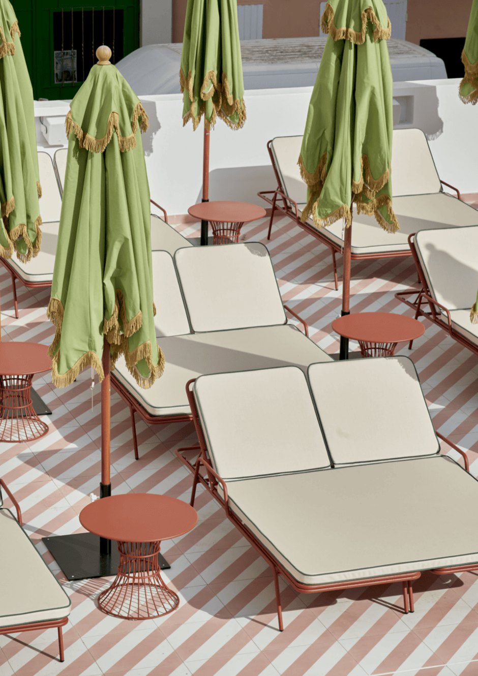 Terracotta-framed sunloungers with cream mattresses and small terracotta pedestal side tables on pink-and-white striped tiling, green fringed parasols furled above.