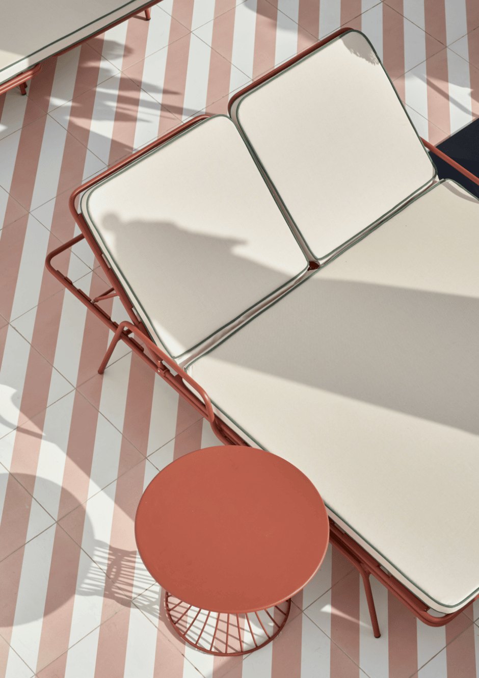 Seen from above, a cream-cushioned sunlounger and a round terracotta side table casting long shadows across striped tiling.