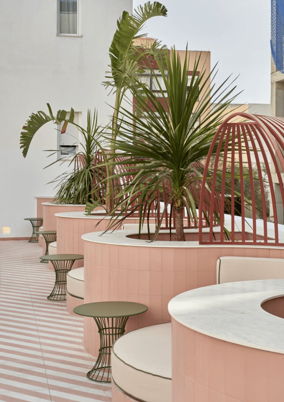 Palms in a pale pink planter behind curved terracotta screens, with green and terracotta pedestal tables beside a curved built-in bench.