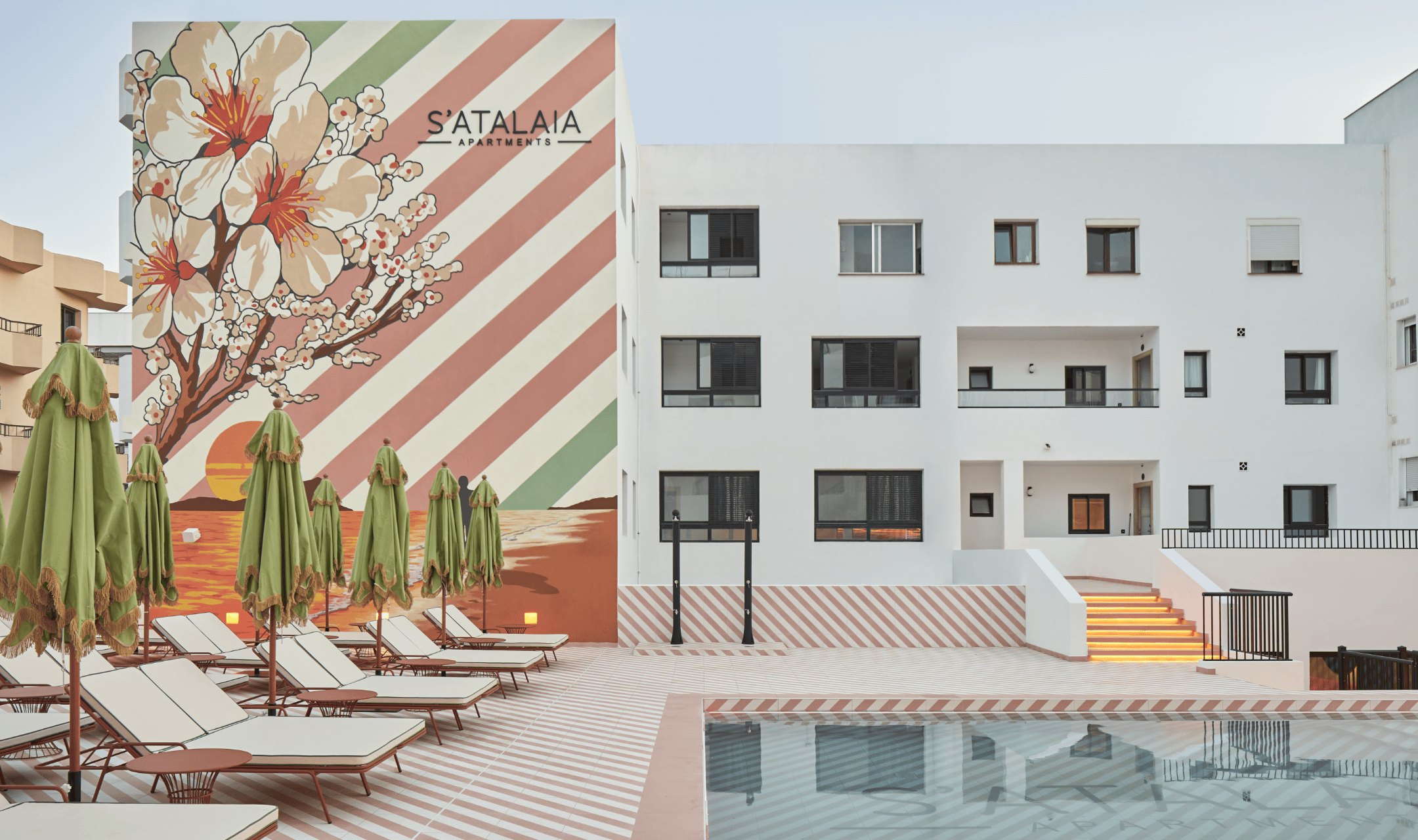 The pool courtyard at S’Atalaia — a painted almond-blossom mural and pink-and-white striped rendering behind a row of terracotta-framed sunloungers and furled green parasols along the water.