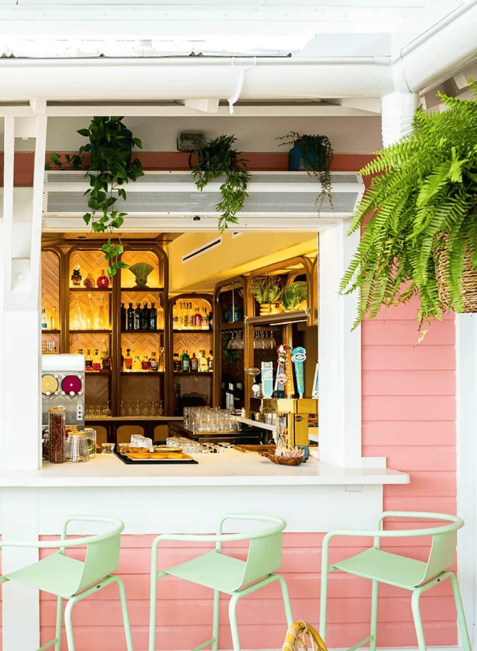 A close view of the bar hatch — coral clapboard, ferns overhead, three mint-green stools drawn up to the counter.
