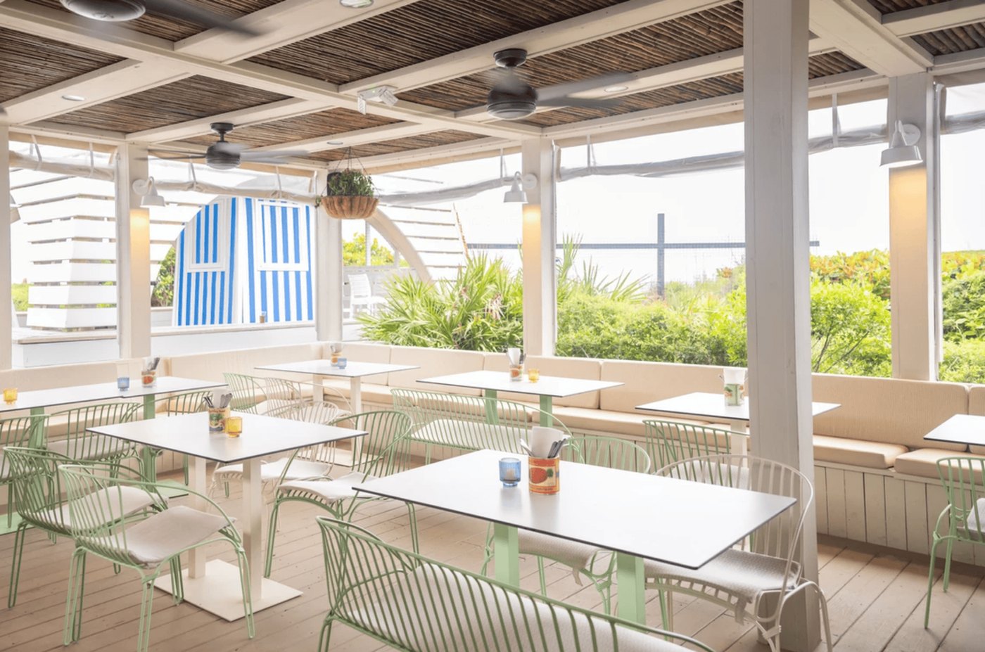 The covered deck: mint-green chairs at white tables under a slatted ceiling, a built-in bench along the wall and a blue-and-white striped cabana beyond.