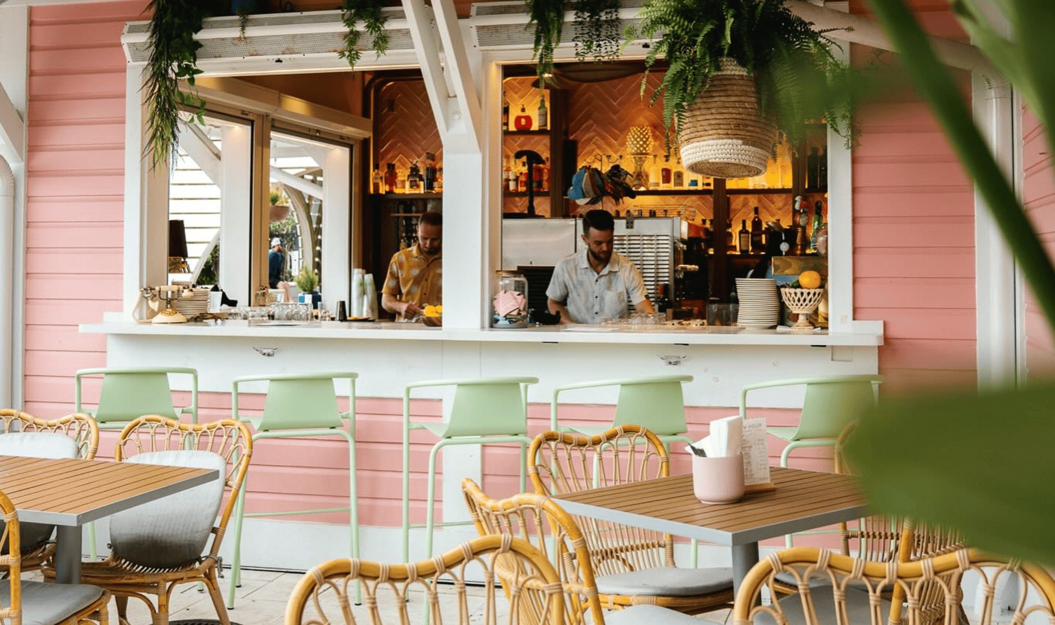 The Daytrader bar — a coral-pink clapboard servery with mint-green stools along the counter, rattan armchairs and timber-topped tables in front.