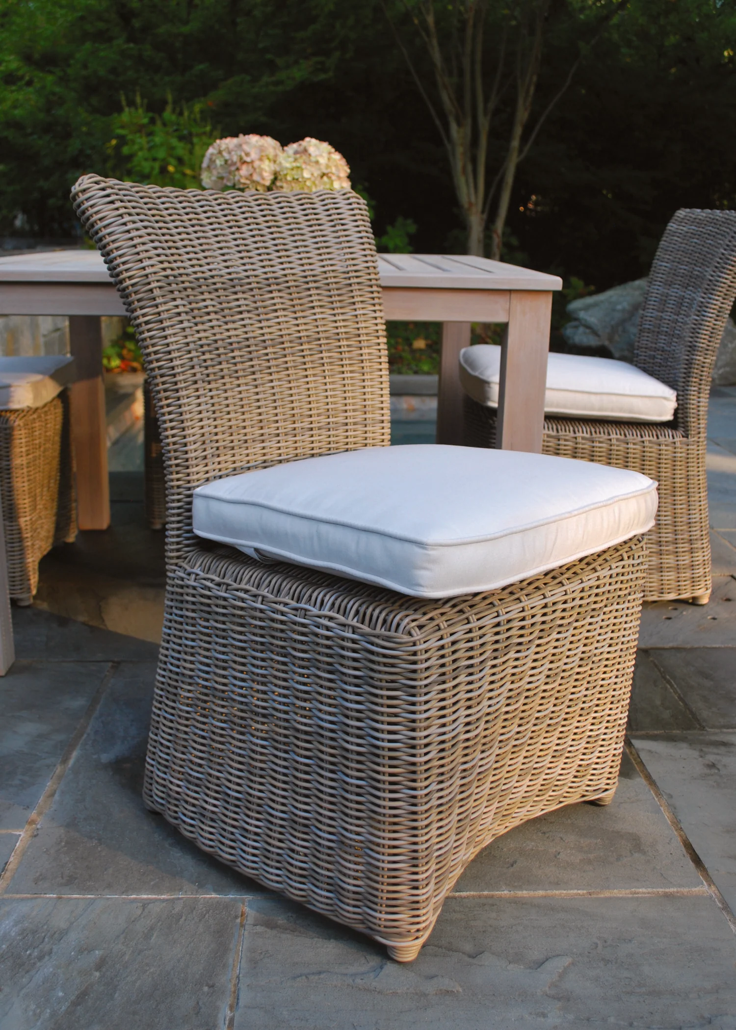 Sag Harbor — Woven dining chairs with white seat cushions drawn up to a pale teak table on bluestone paving at dusk.