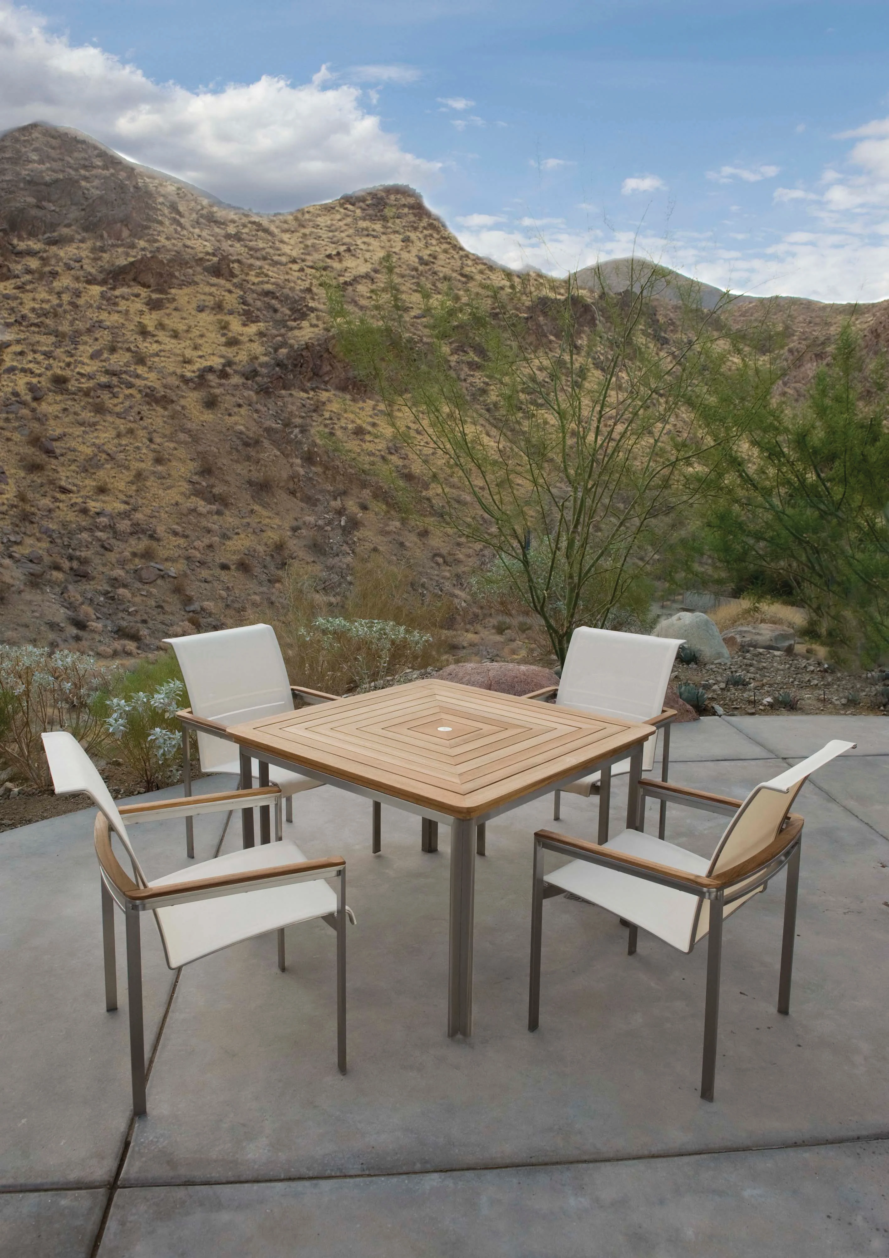 Tivoli — Square teak-topped dining table and four stainless sling armchairs on a poured concrete terrace below a desert hillside.