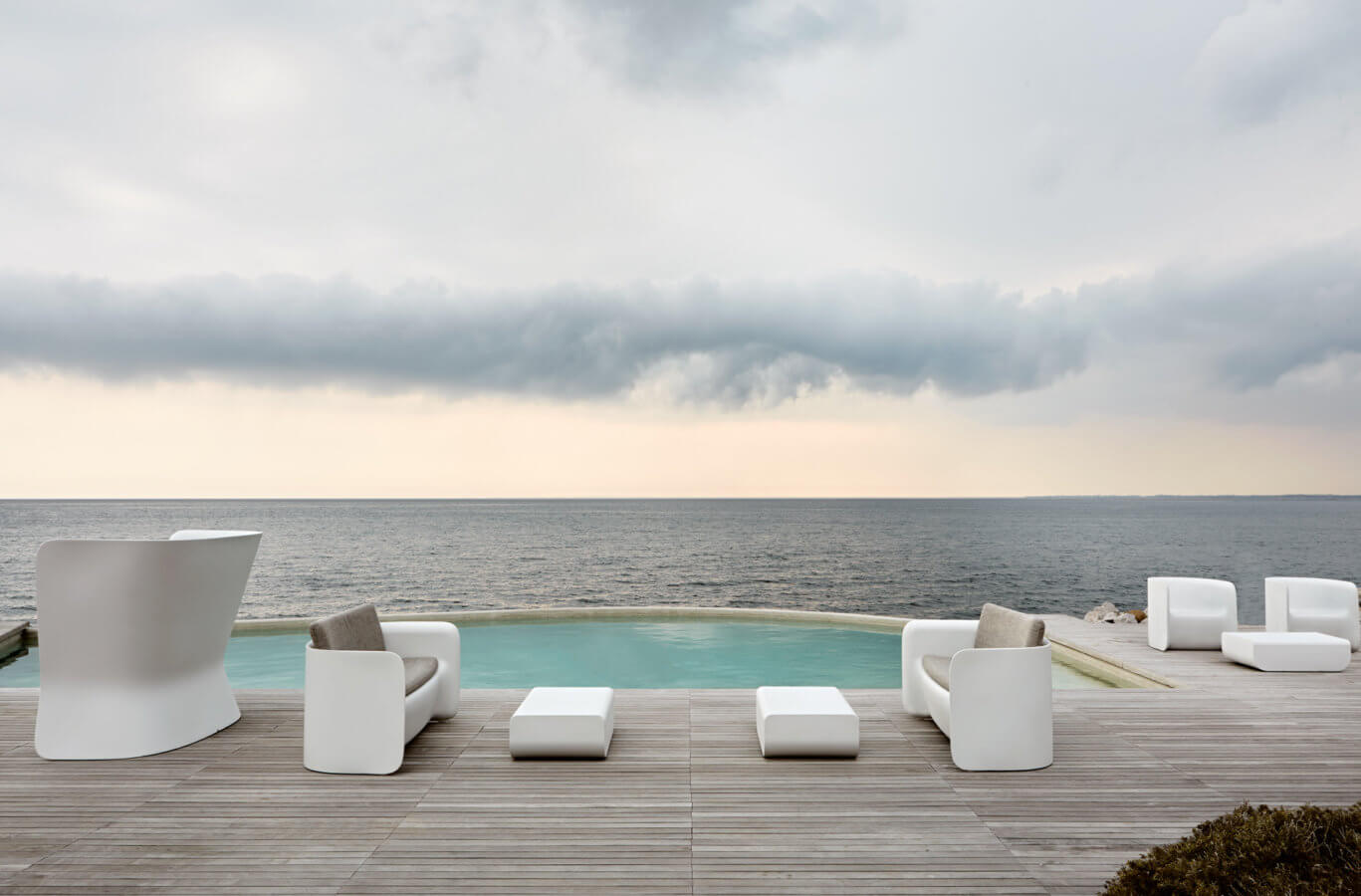 White Nova S sofa sections and 2054 coffee tables on timber decking above an infinity pool, the sea beyond