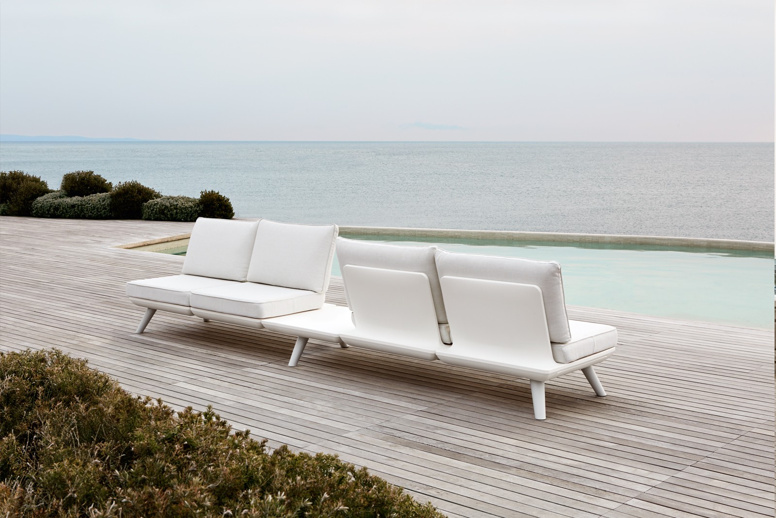 A white Samurai sofa, two armless sections set back to back, on decking beside an infinity pool and the sea