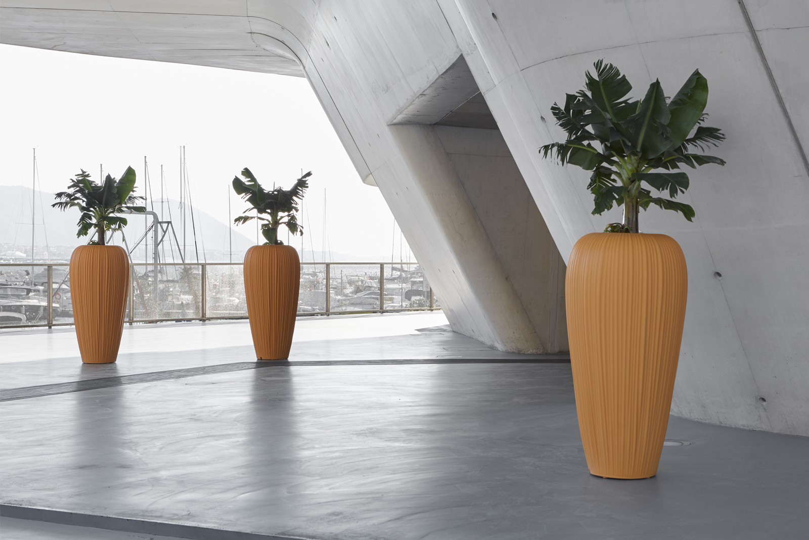 Tall Skins pot holders in banana yellow, planted with banana palms, beneath a concrete portico beside a marina