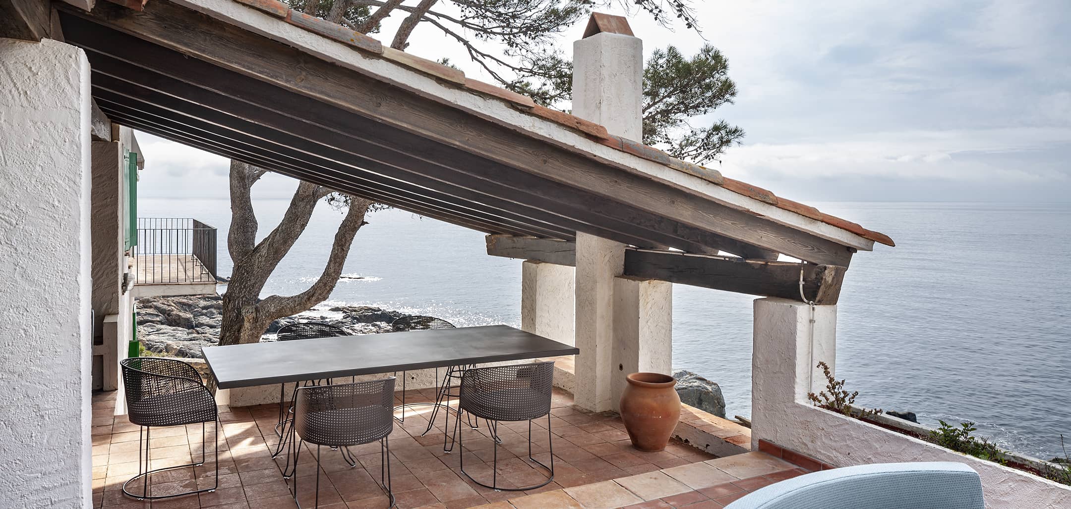 Arena dining chairs in perforated steel around a Capri table on a sea terrace, under a timber pergola with a terracotta tiled floor — iSiMAR catalogue photography