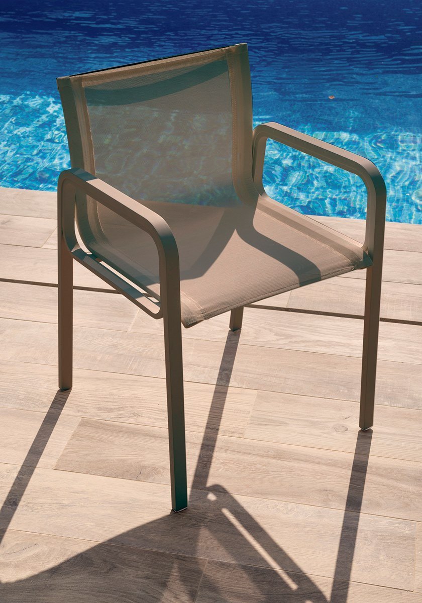 A taupe sling-mesh dining armchair on a timber pool deck, shot close and angled down, with bright blue pool water filling the background.