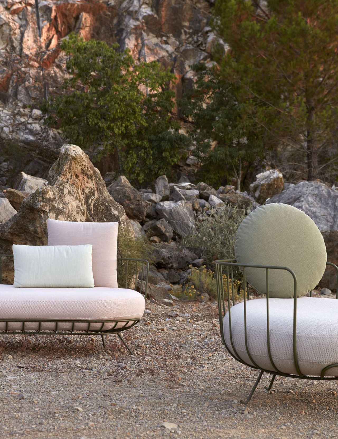 A wire-frame daybed with pink and white cushions and a round-backed wire-frame lounge chair in sage, set on gravel before a scrubby Mediterranean hillside.