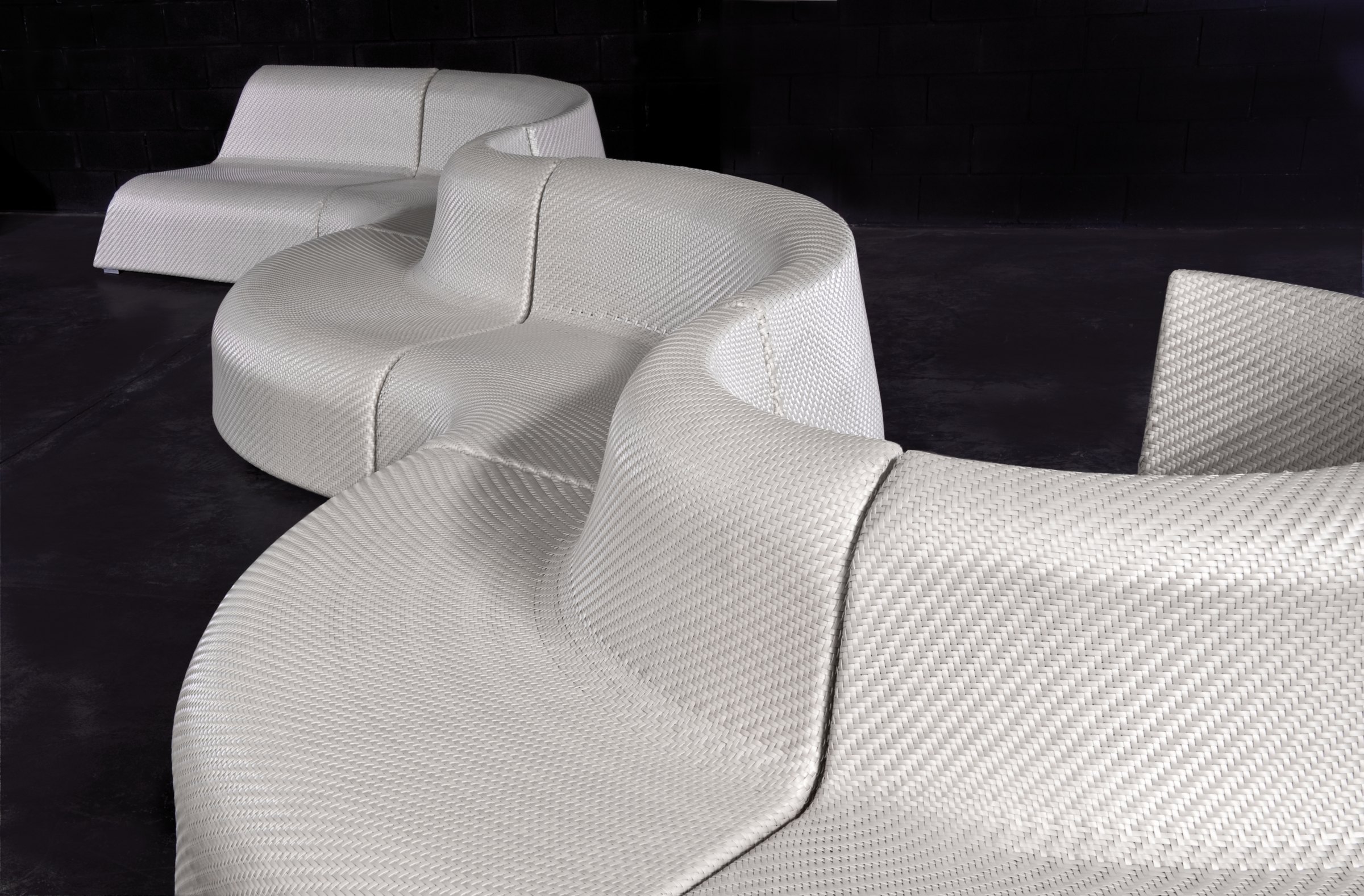 Three white woven bench modules in sinuous wave forms, shown close and receding into a dark gallery-lit room with a black brick wall.