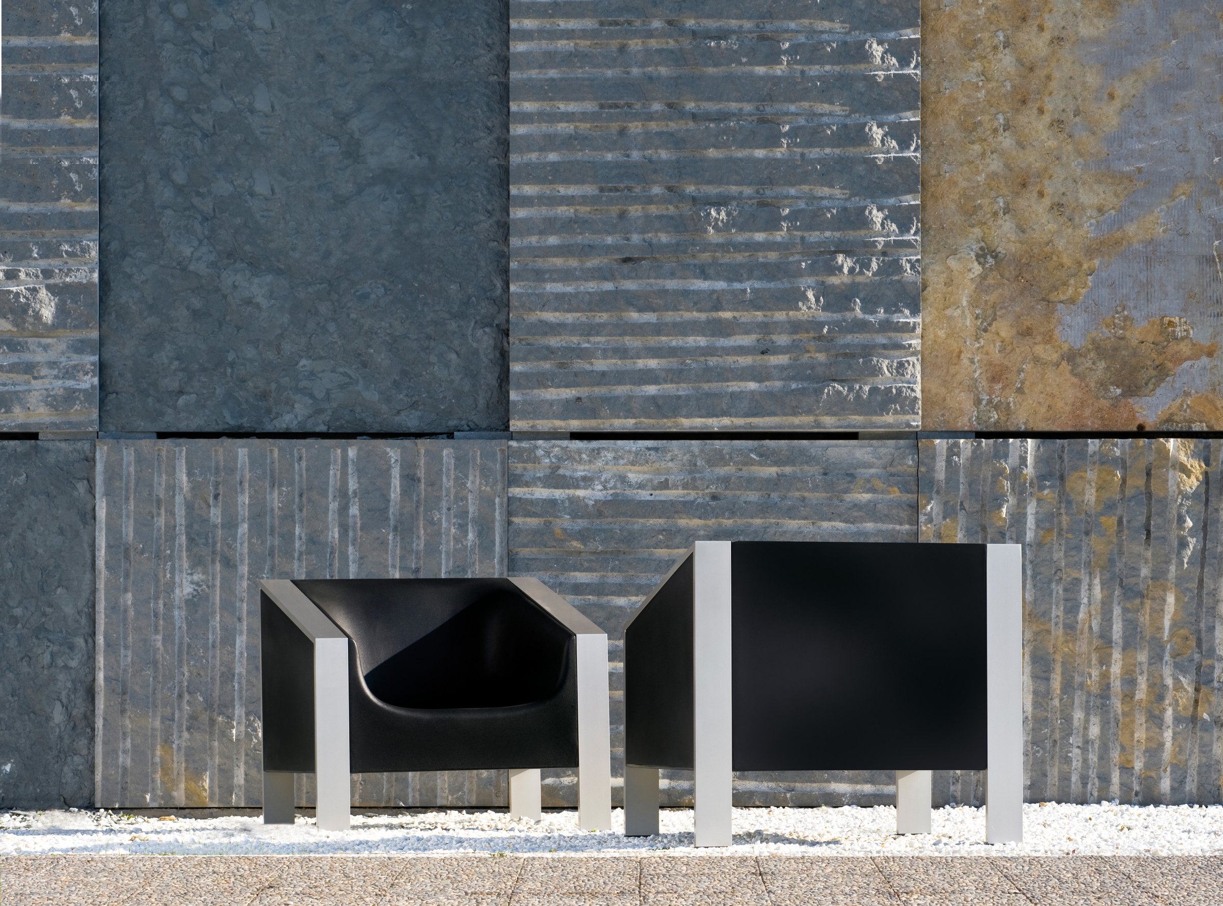 Two black cube-framed armchairs on brushed steel legs, standing on white gravel against a wall of dark slate and rust-toned stone tiles.