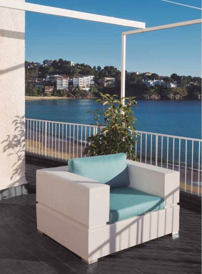 A white cube-framed armchair with an aqua cushion and bolster on a balcony, a small potted shrub beside it and a Mediterranean bay with a white hillside town beyond the railing.