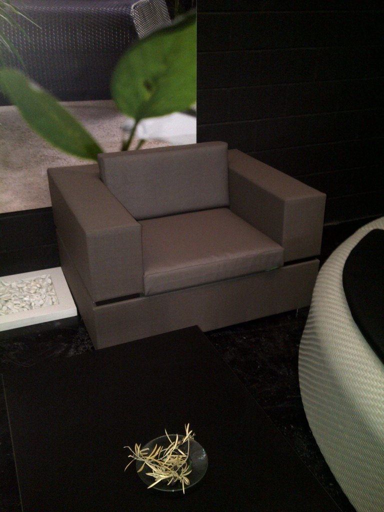 A taupe sling-fabric armchair from the Cubic Sling range shown at a trade display, with a potted plant, a dish of rosemary sprigs and a photo backdrop behind it.