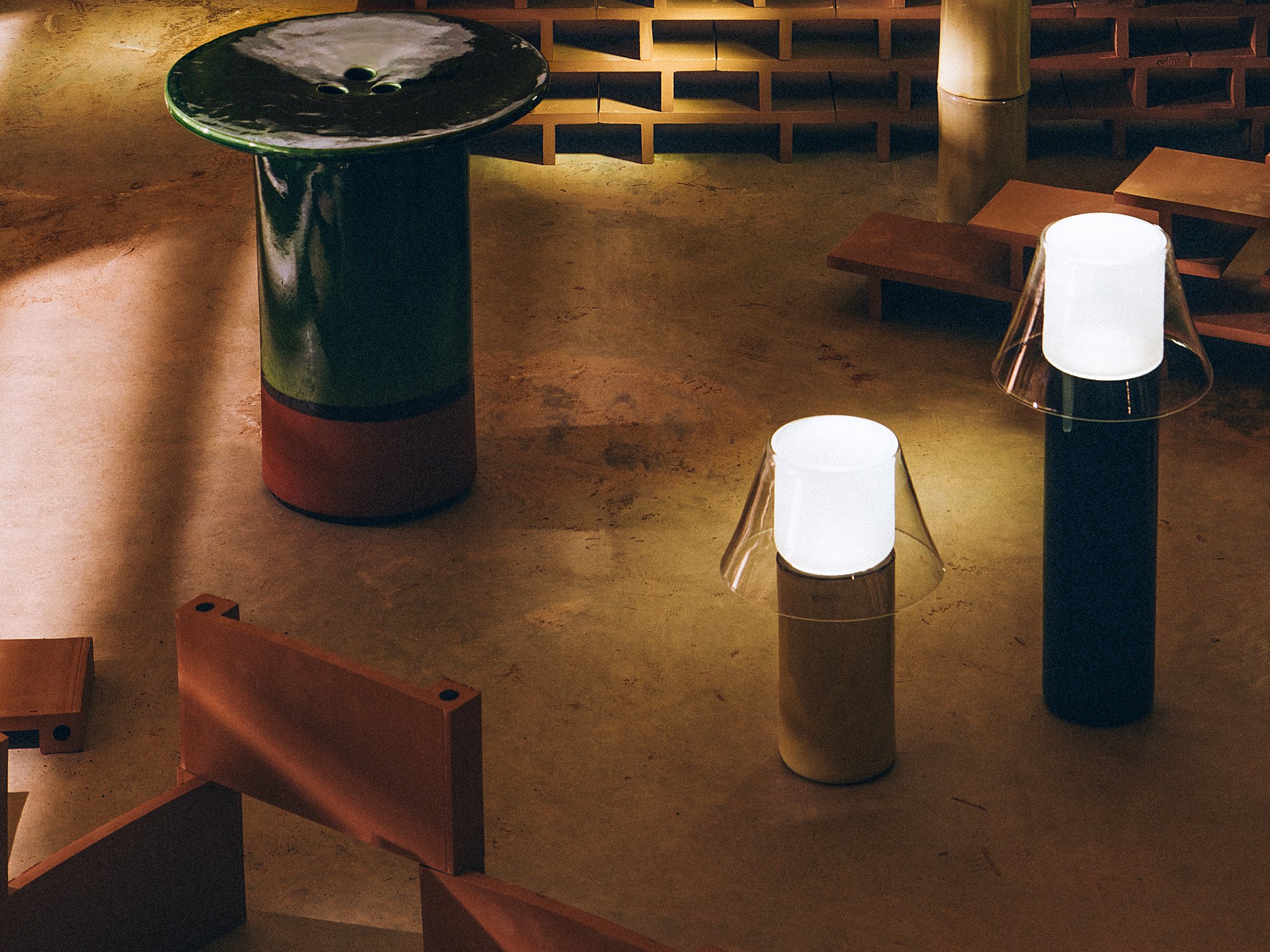 A glazed ceramic Tub side table in green-to-rust tones beside two glowing cordless lamps with conical glass shades, standing on a concrete floor before a dim, terracotta brick lattice wall.