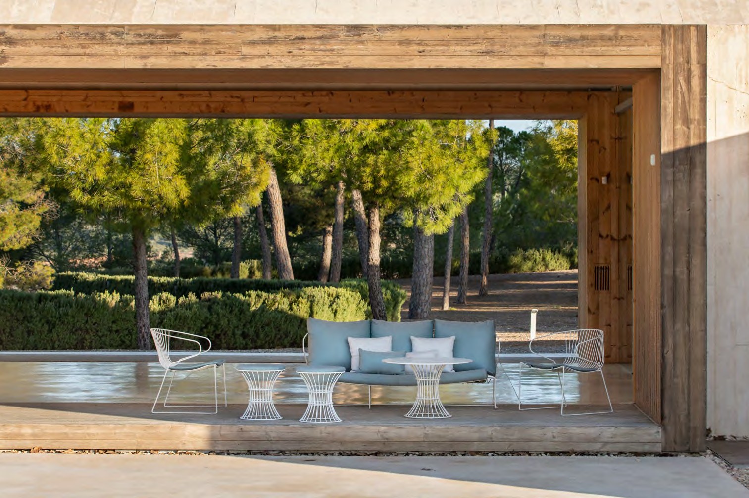 A duck-egg blue sofa and two armchairs in white galvanised wire, under a timber pavilion beside a pool ringed with pines — iSiMAR catalogue photography